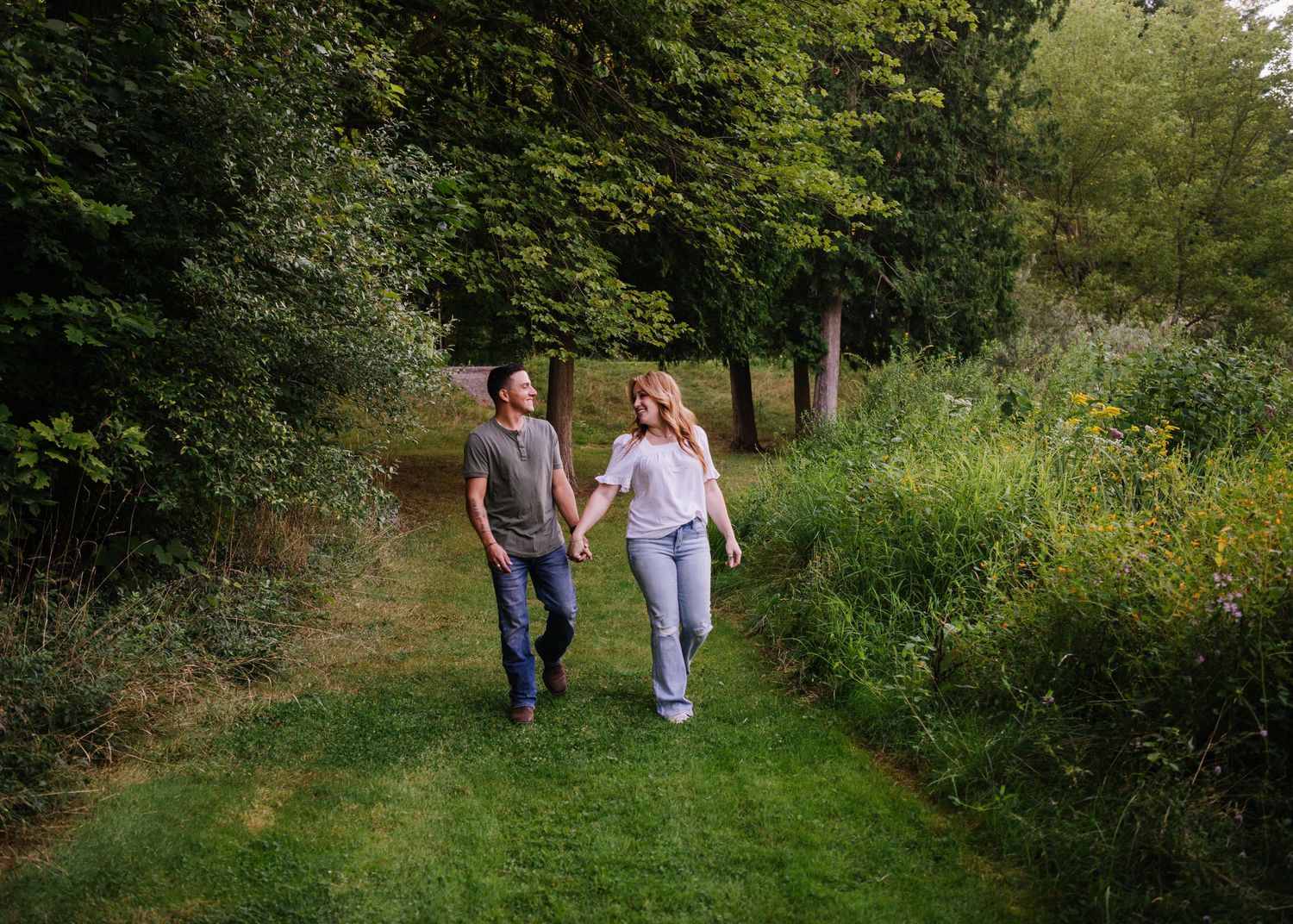 A couple walks hand in hand along a winding nature trail surrounded by lush green foliage on a sunny day.