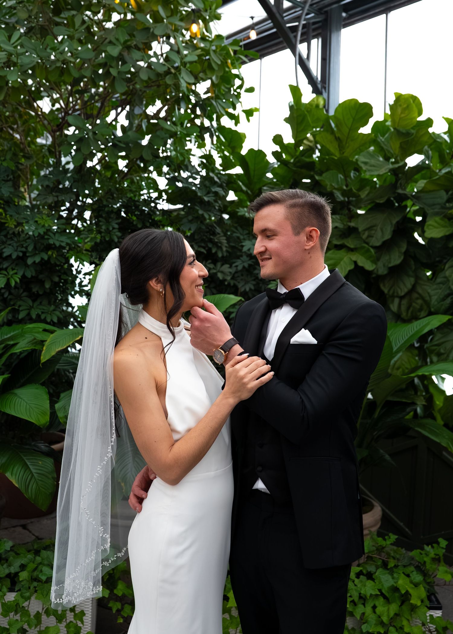 Bride and groom share a romantic moment surrounded by lush greenery, she in a white halter gown with veil, he in a black tuxedo.