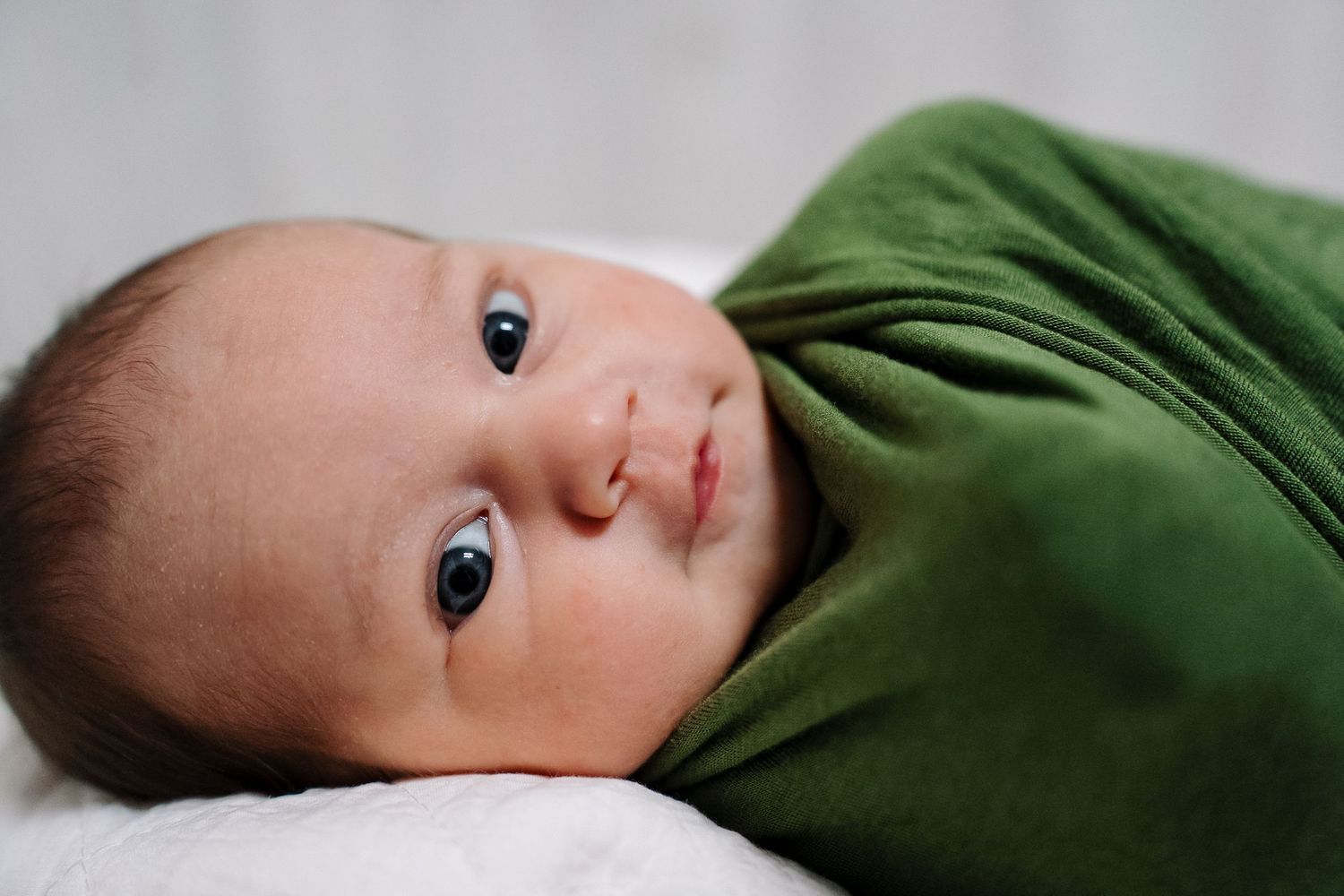 Newborn baby swaddled in green blanket with blue eyes lying on white surface.