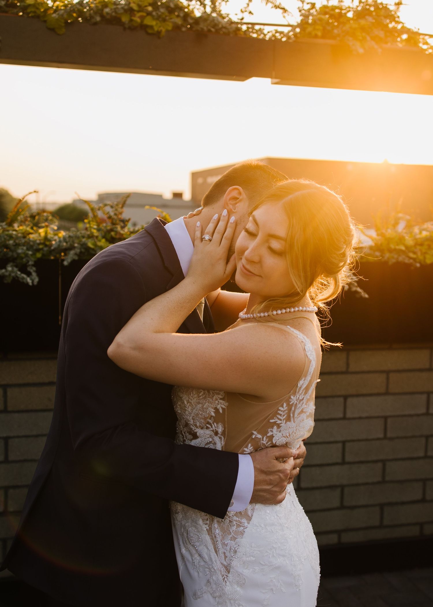 Romantic wedding couple embraces during golden hour sunset with dramatic backlighting against brick wall.
