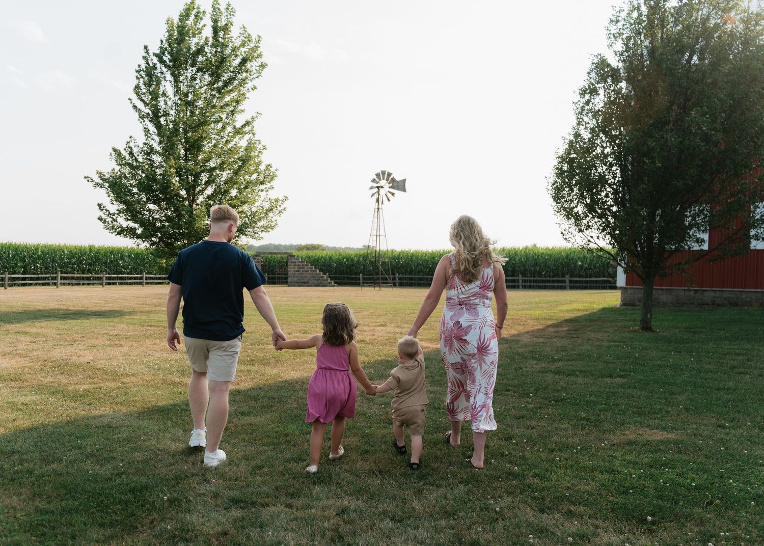 Family of four walking hand-in-hand through a farm field with windmill and cornfield in background.