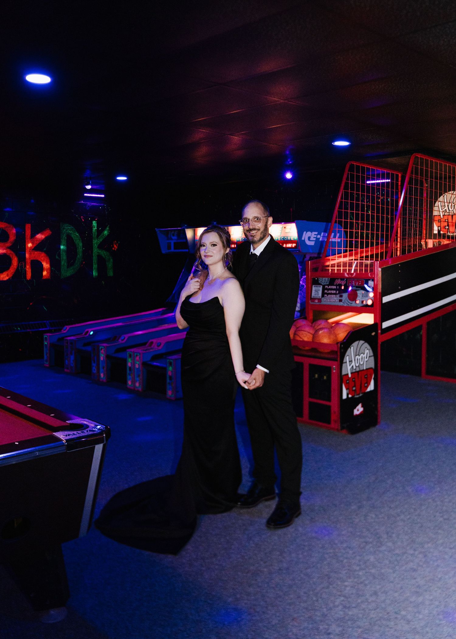 A couple poses in formal attire at a neon-lit arcade or entertainment venue with pool tables and gaming equipment.