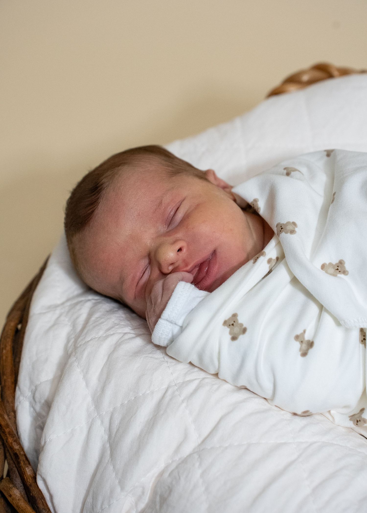 Newborn baby sleeping peacefully in a wicker basket, wrapped in a white outfit with small bear prints.