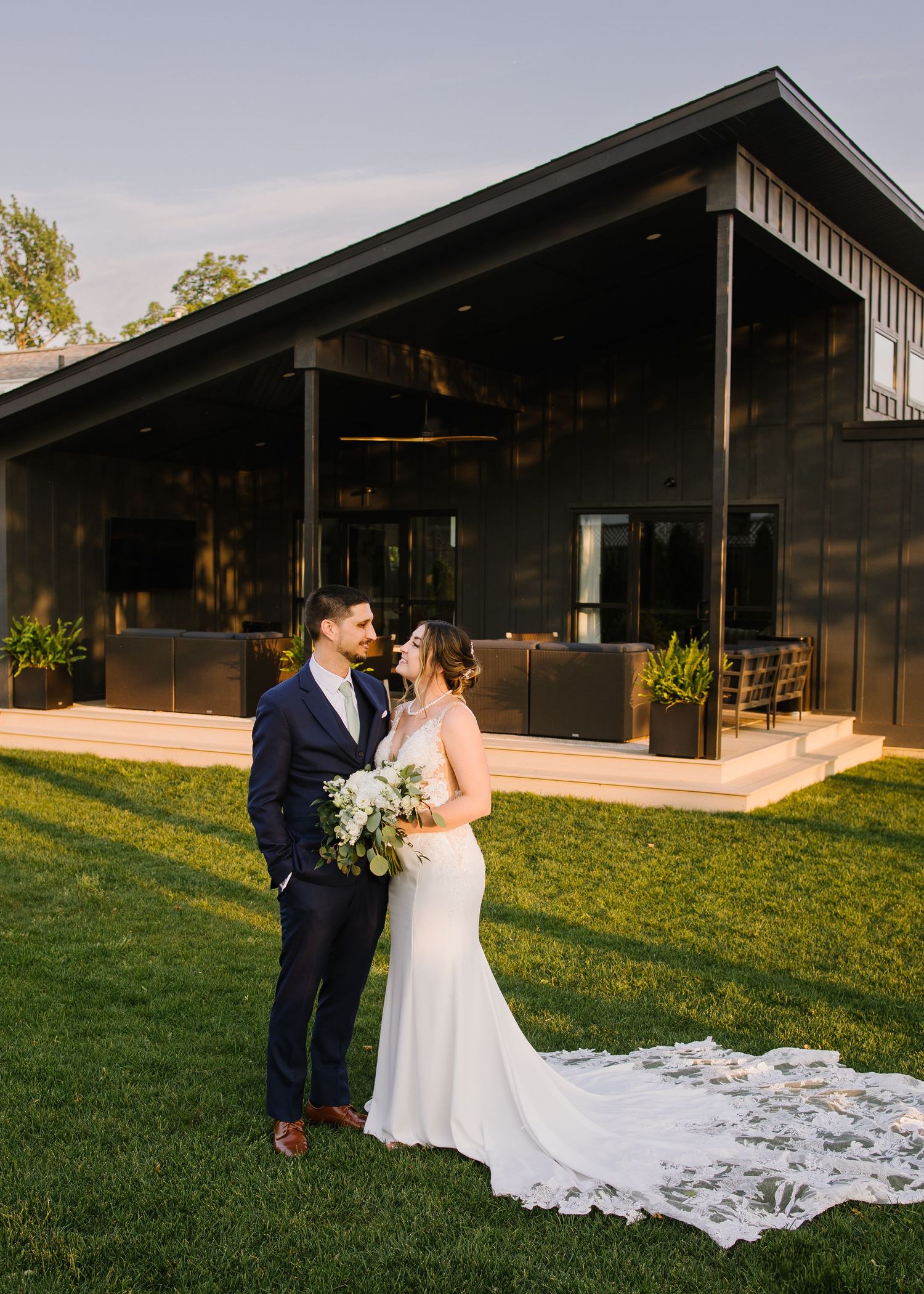 Wedding couple shares romantic moments at twilight on a grassy lawn beside a modern black barn venue.