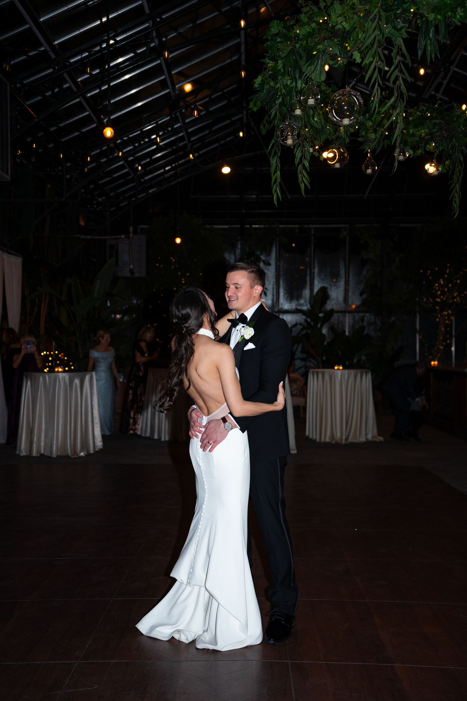 Bride and groom share their first dance in an elegant greenhouse wedding venue with string lights and lush greenery overhead.