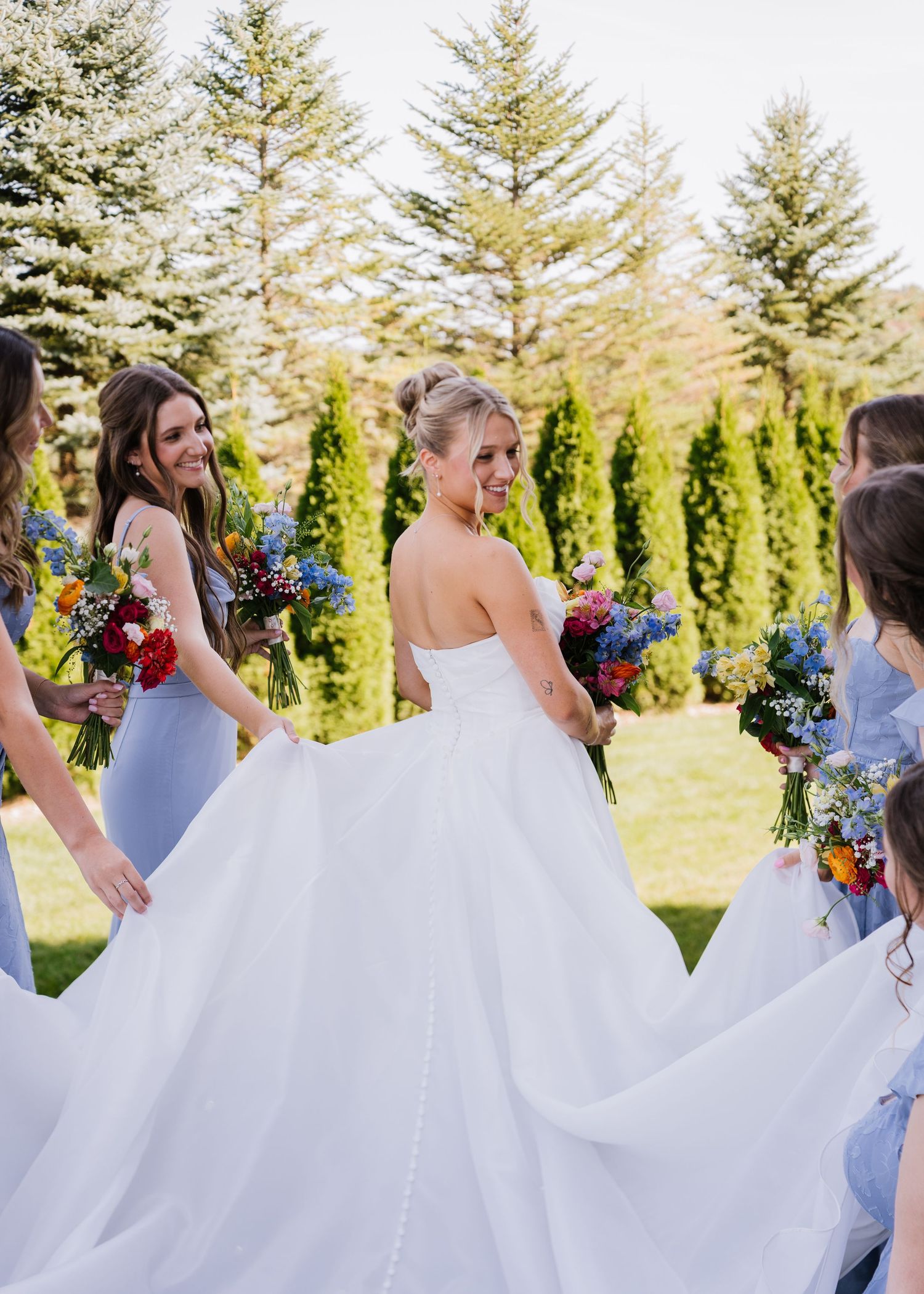 Bridesmaids in white dresses twirl and dance joyfully with colorful bouquets against an evergreen backdrop.