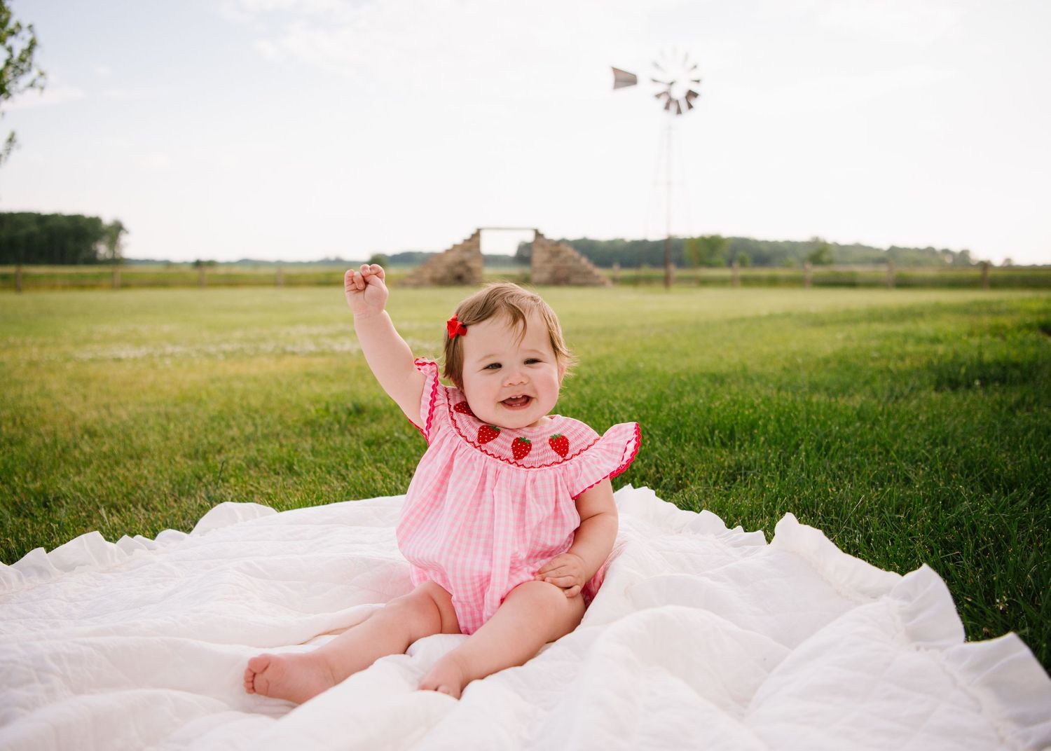 A small child in a pink dress sits on a white blanket in a grassy field on a sunny day.