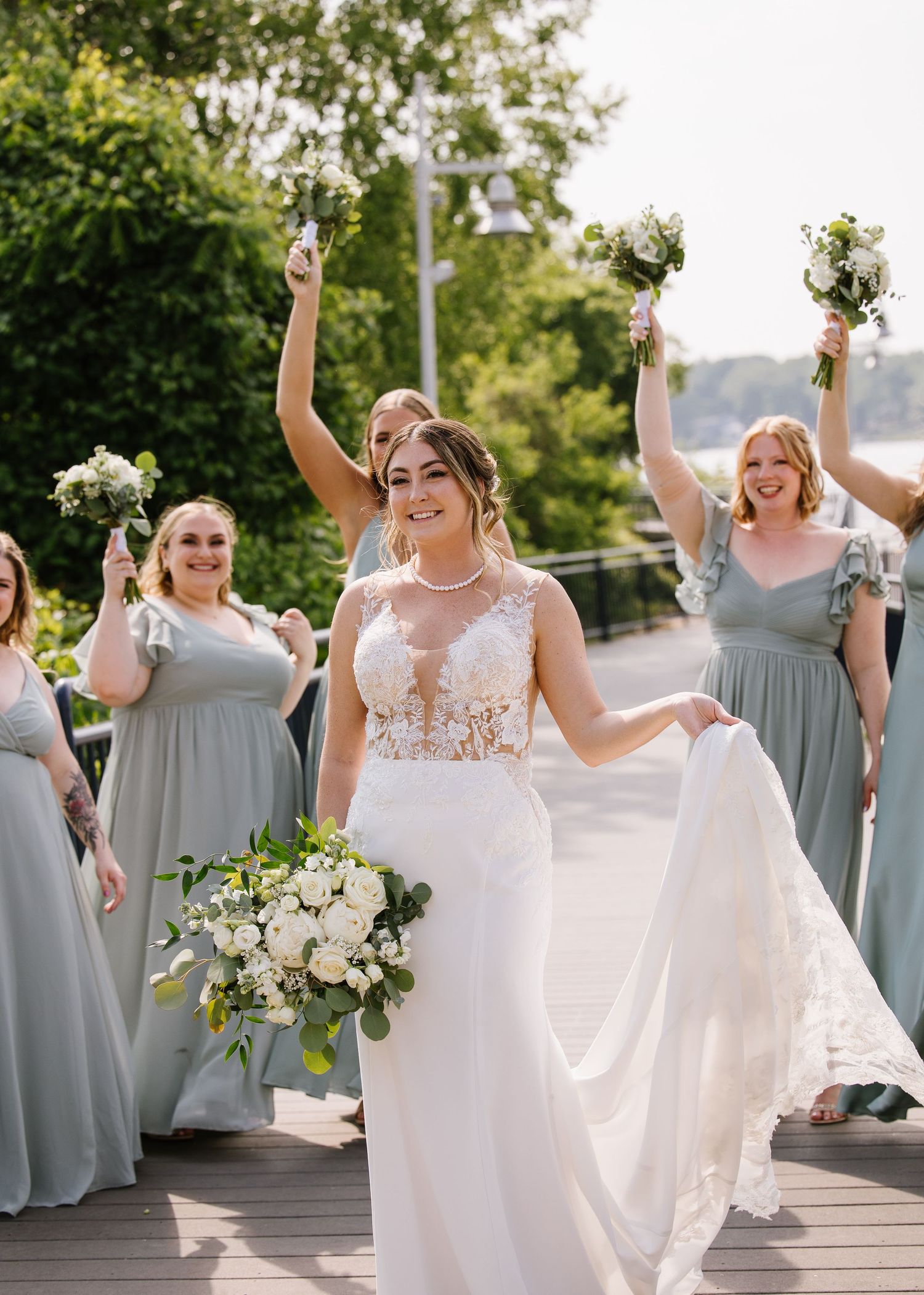 Bridesmaids in flowing sage green dresses celebrate joyfully on a wooden deck with white floral bouquets.