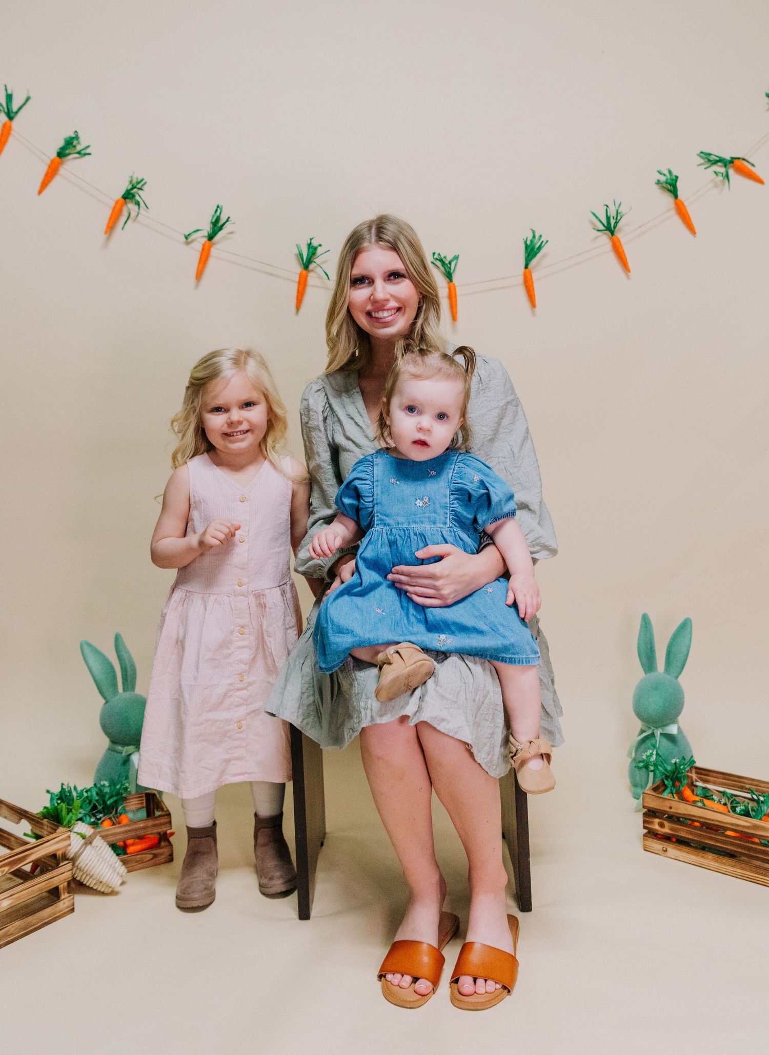 A spring photo setup with carrot garland and green bunny decorations against a beige wall.
