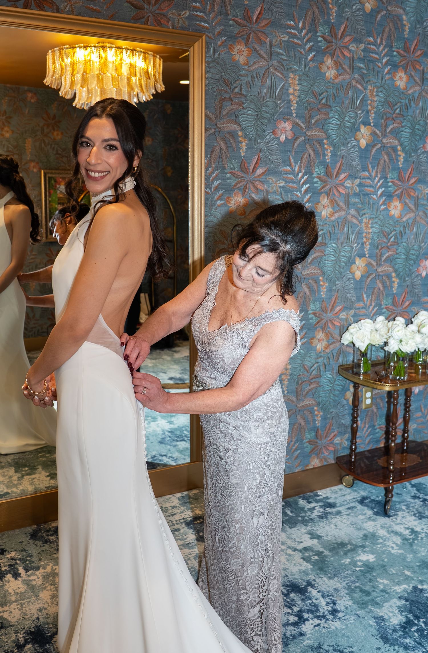 A bride in a white gown smiles as an older woman in silver lace helps fasten her dress in an elegant floral room.