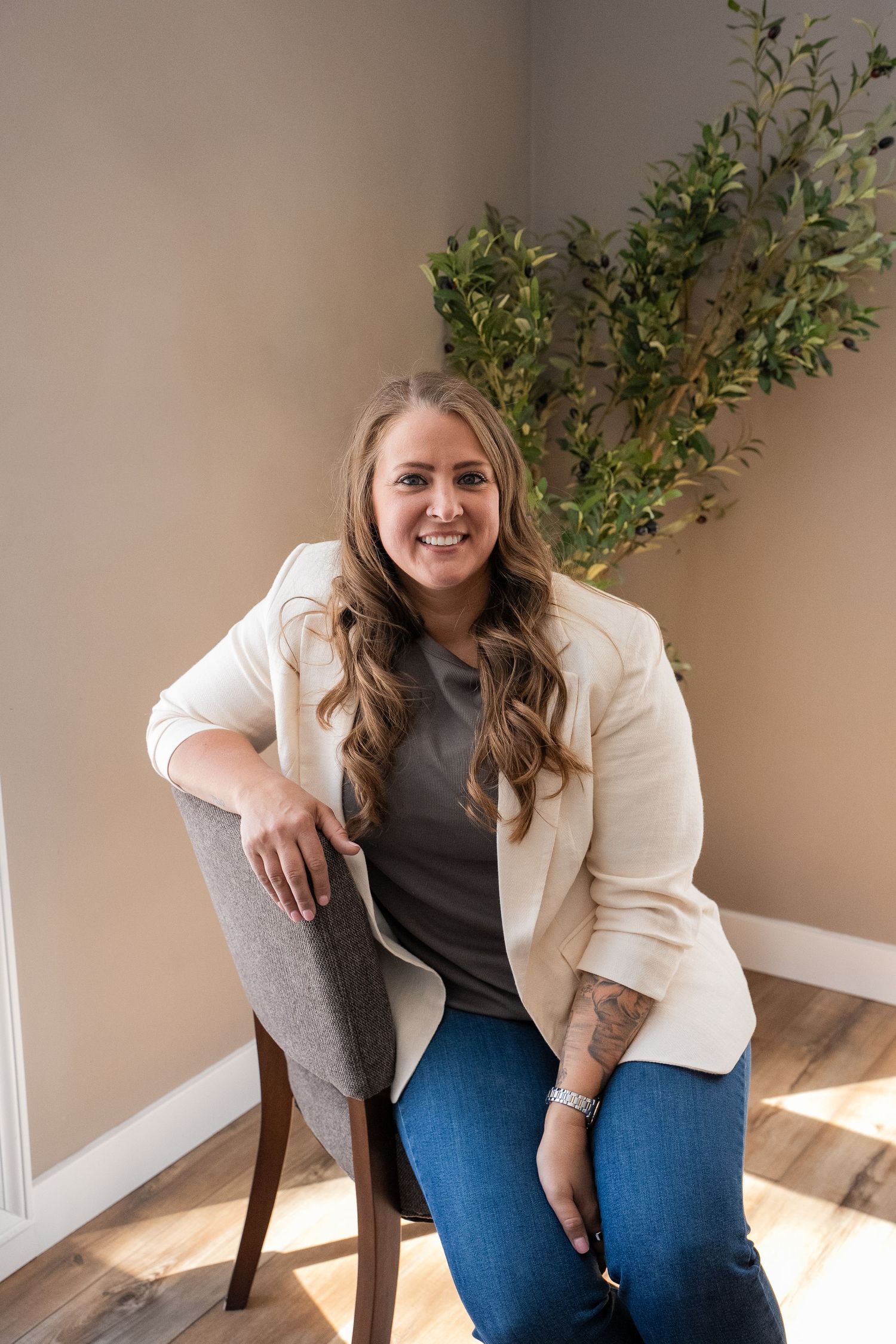 Smiling woman in white blazer and jeans sits on chair beside green plant in bright neutral room.