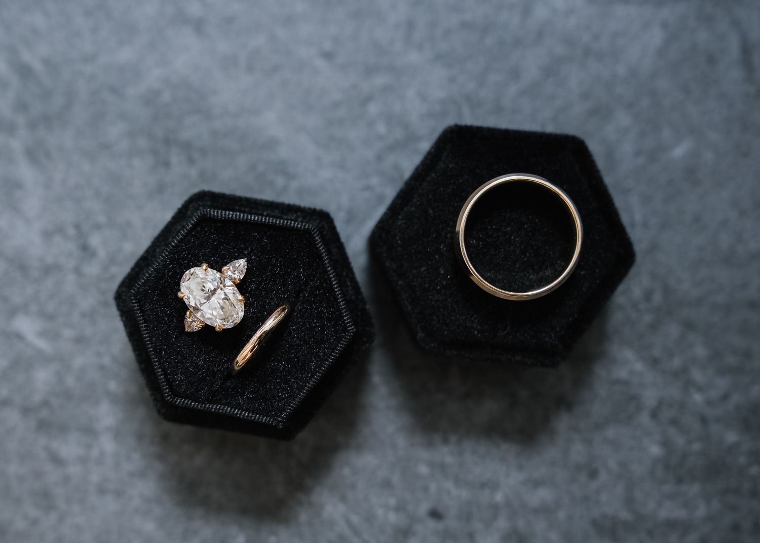 Diamond engagement ring and gold wedding band displayed in black velvet hexagon ring boxes on gray surface.