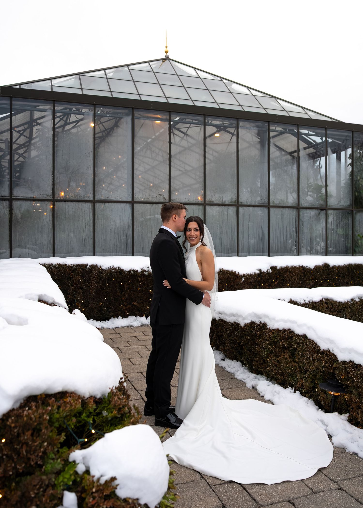 Bride and groom embrace in snow-covered garden outside glass greenhouse conservatory during winter wedding.