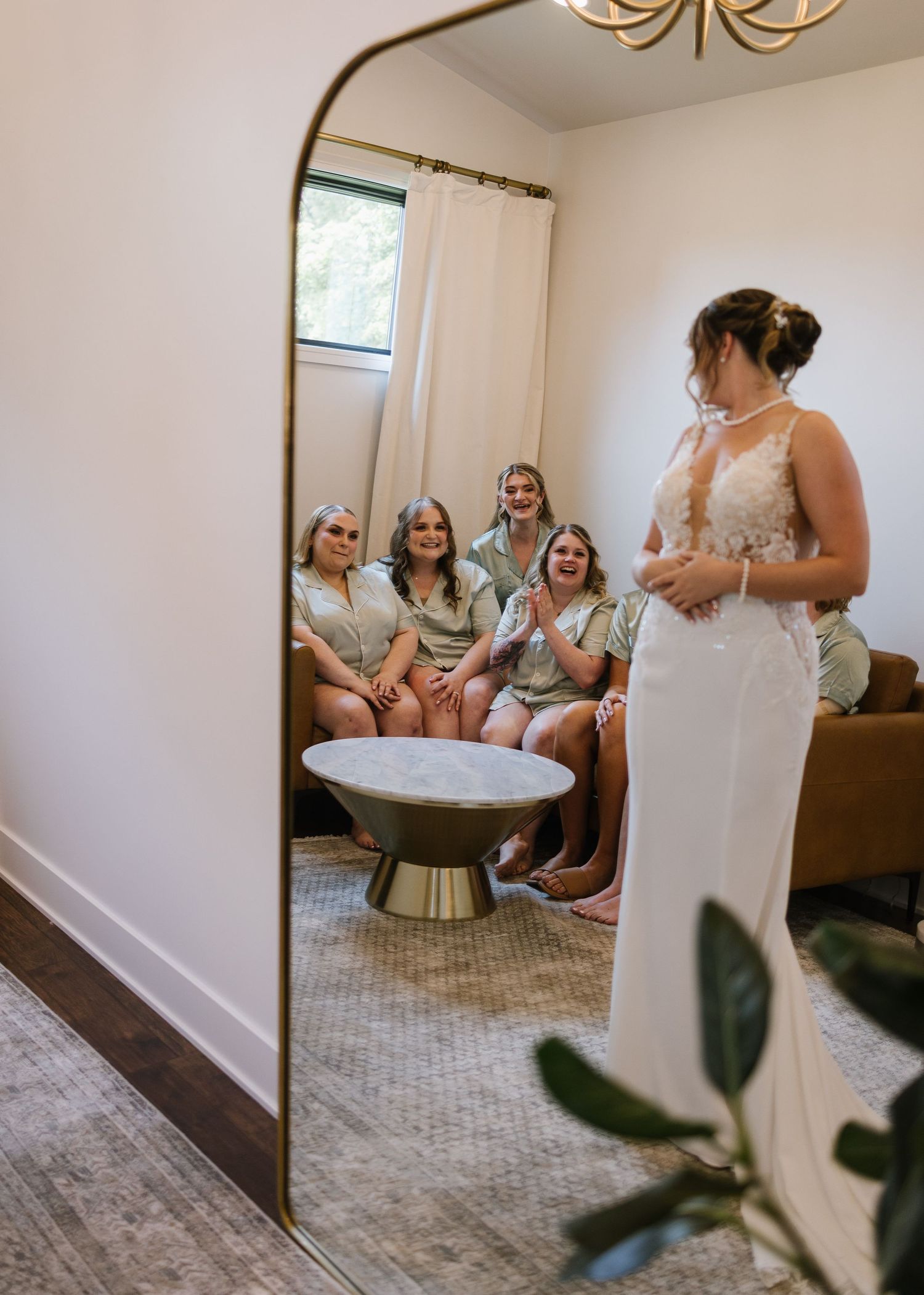 A bride in a white fitted dress stands by a mirror while bridesmaids in mint dresses sit on a couch in the reflection.