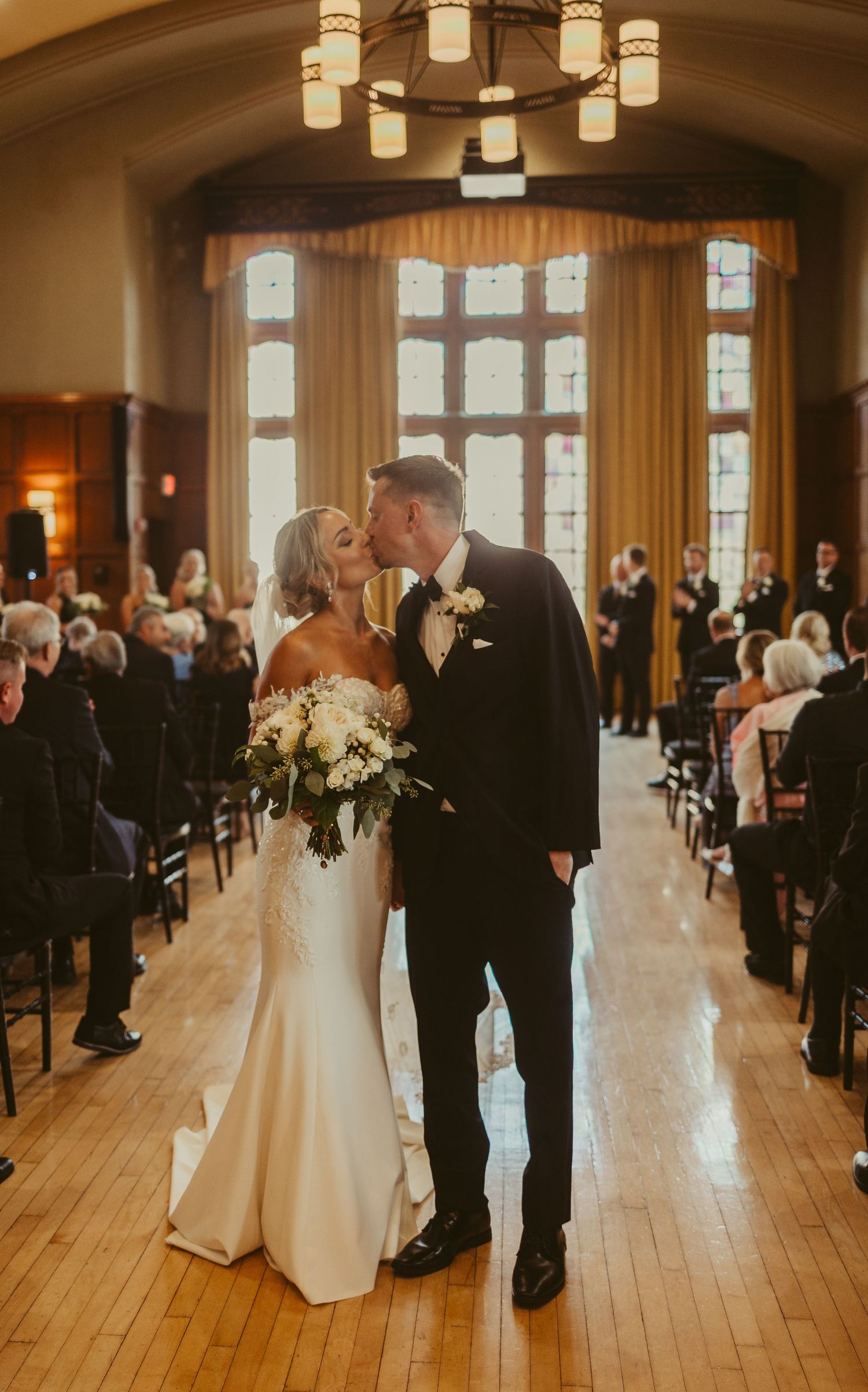A romantic wedding ceremony scene in an elegant venue with hardwood floors and tall windows, illuminated by warm lighting.