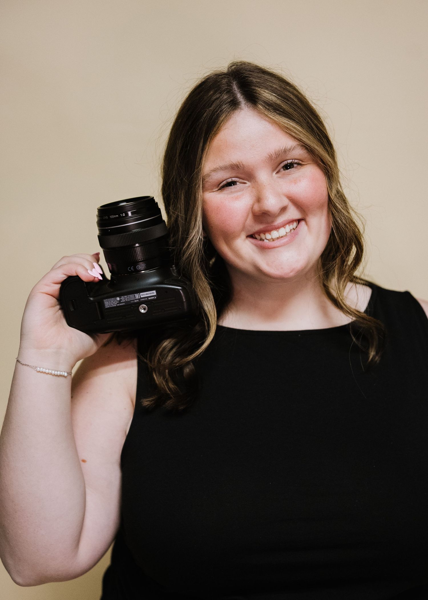 Young woman smiling and holding a DSLR camera against a beige background, wearing a black top.