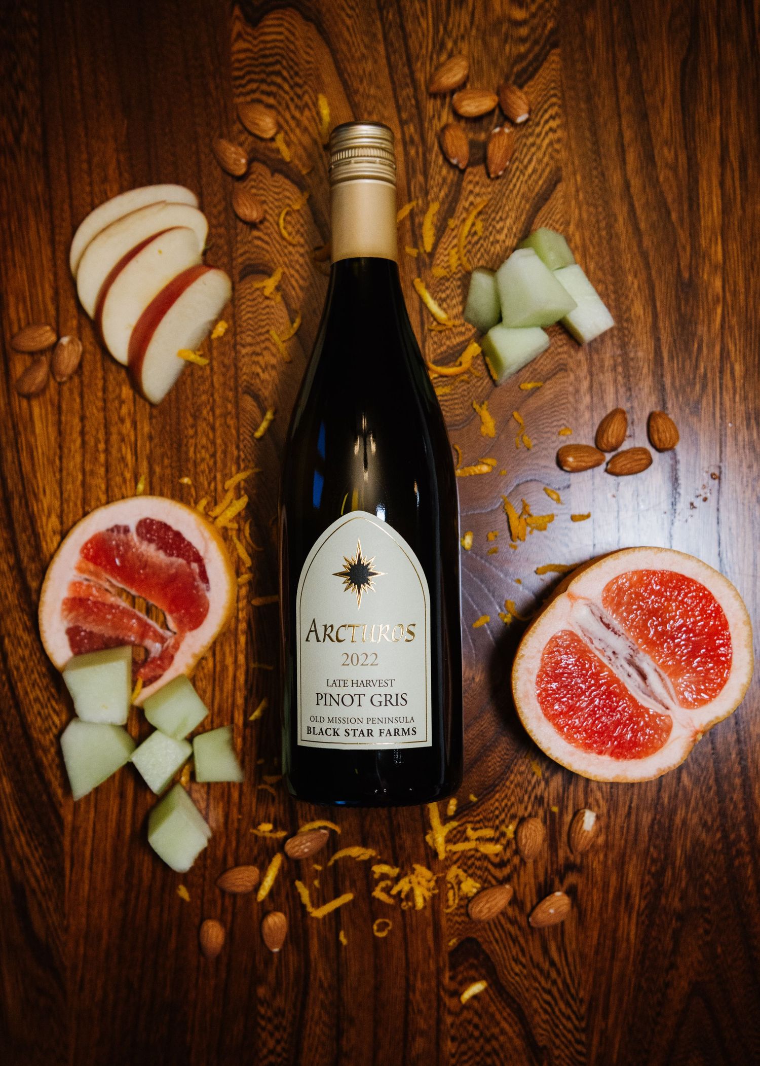 Wine bottle surrounded by sliced fresh grapefruit, melon cubes, and scattered nuts on a wooden surface.