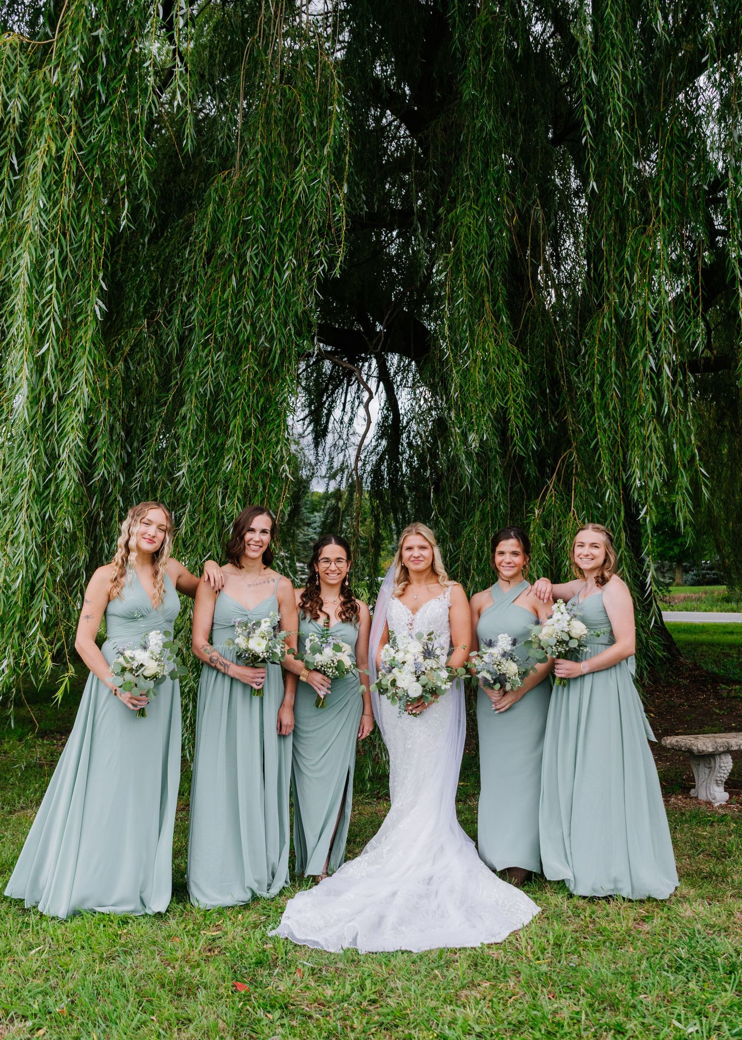 A wedding party in sage green bridesmaid dresses poses under a weeping willow tree on a grassy lawn.