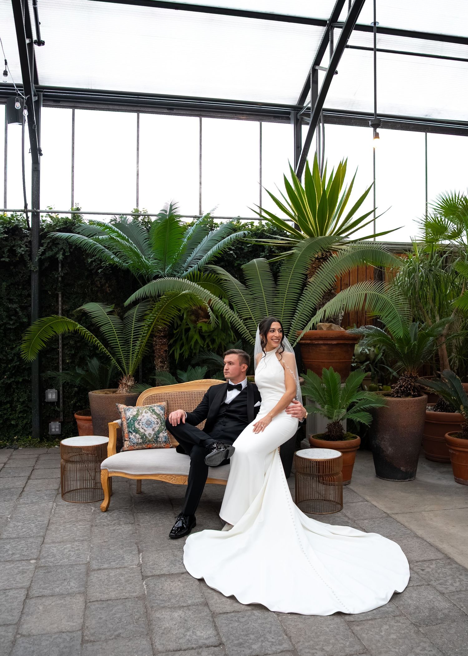 Bride in white gown and groom in tuxedo pose on a vintage sofa in a lush greenhouse botanical garden.
