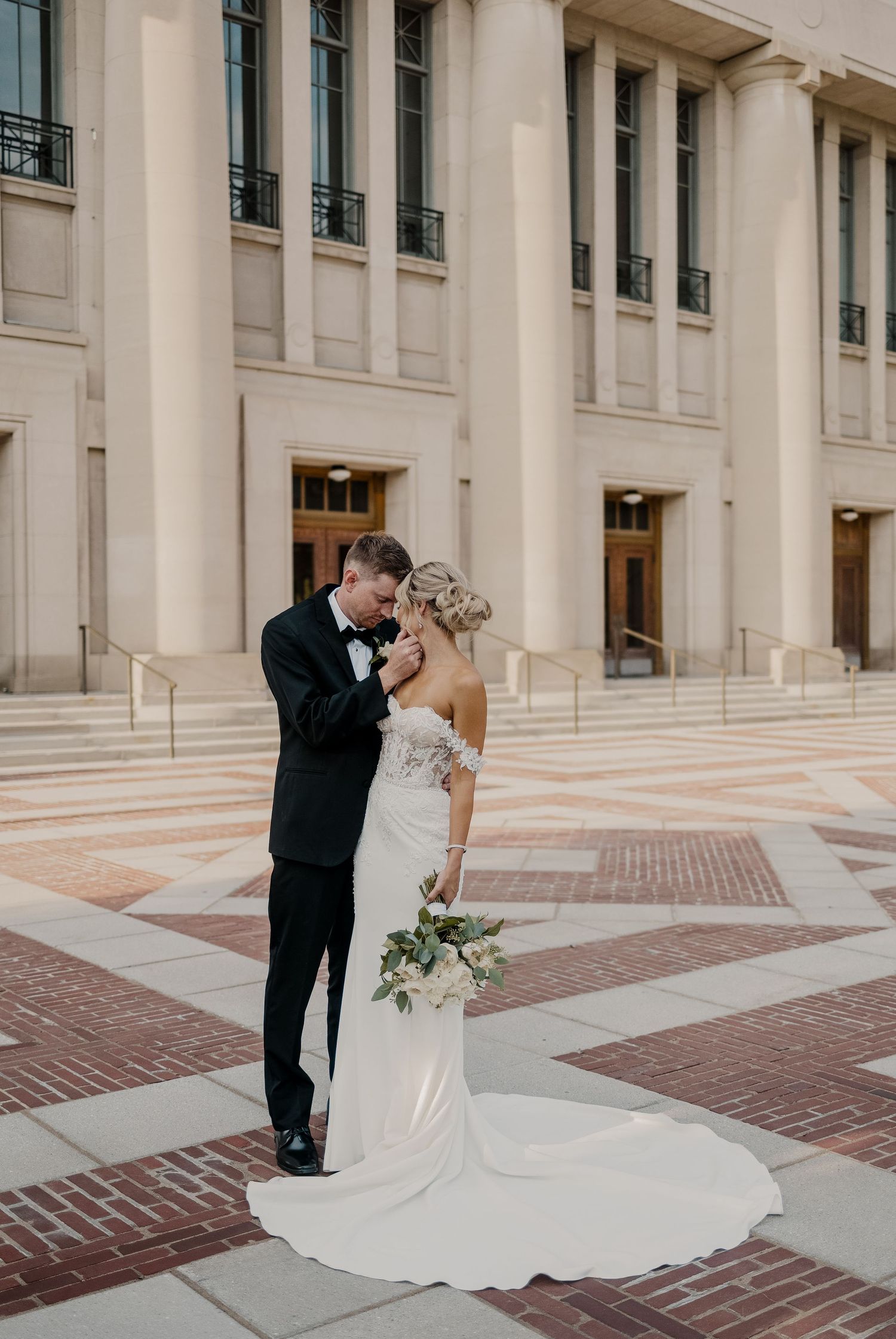 A couple in wedding attire share a romantic moment on elegant stone steps with white columns in the background.