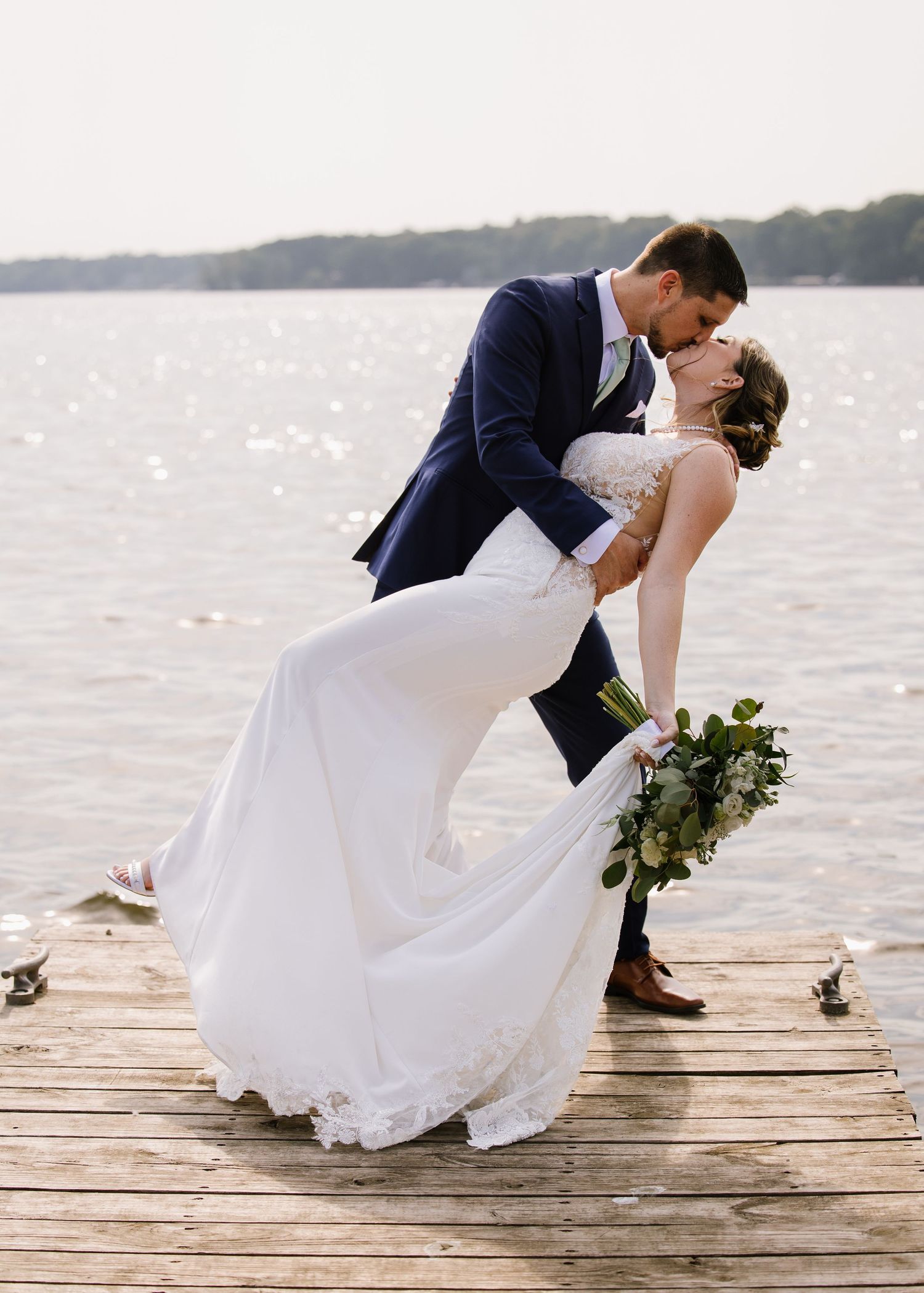 Romantic wedding couple dips in dramatic pose on lakeside dock at sunset creating elegant silhouette against water.