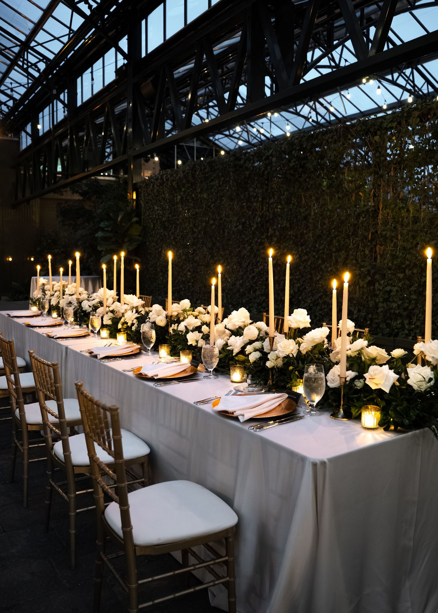 Elegant greenhouse wedding reception with long white table, tall candles, white floral centerpieces, and gold chiavari chairs.