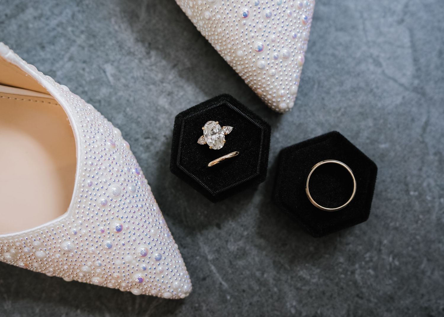 Pearl bridal heels beside a black hexagon velvet ring box holding a diamond engagement ring and gold wedding band.