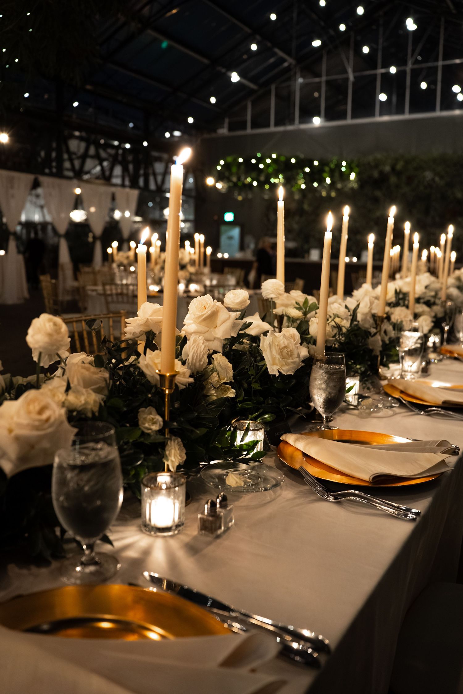 Elegant wedding reception table with white roses, tall gold candles, gold charger plates, and string lights in a dark venue.