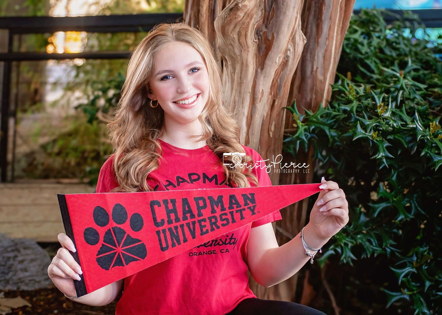 Smiling young woman in red shirt holding a Chapman University pennant outdoors.