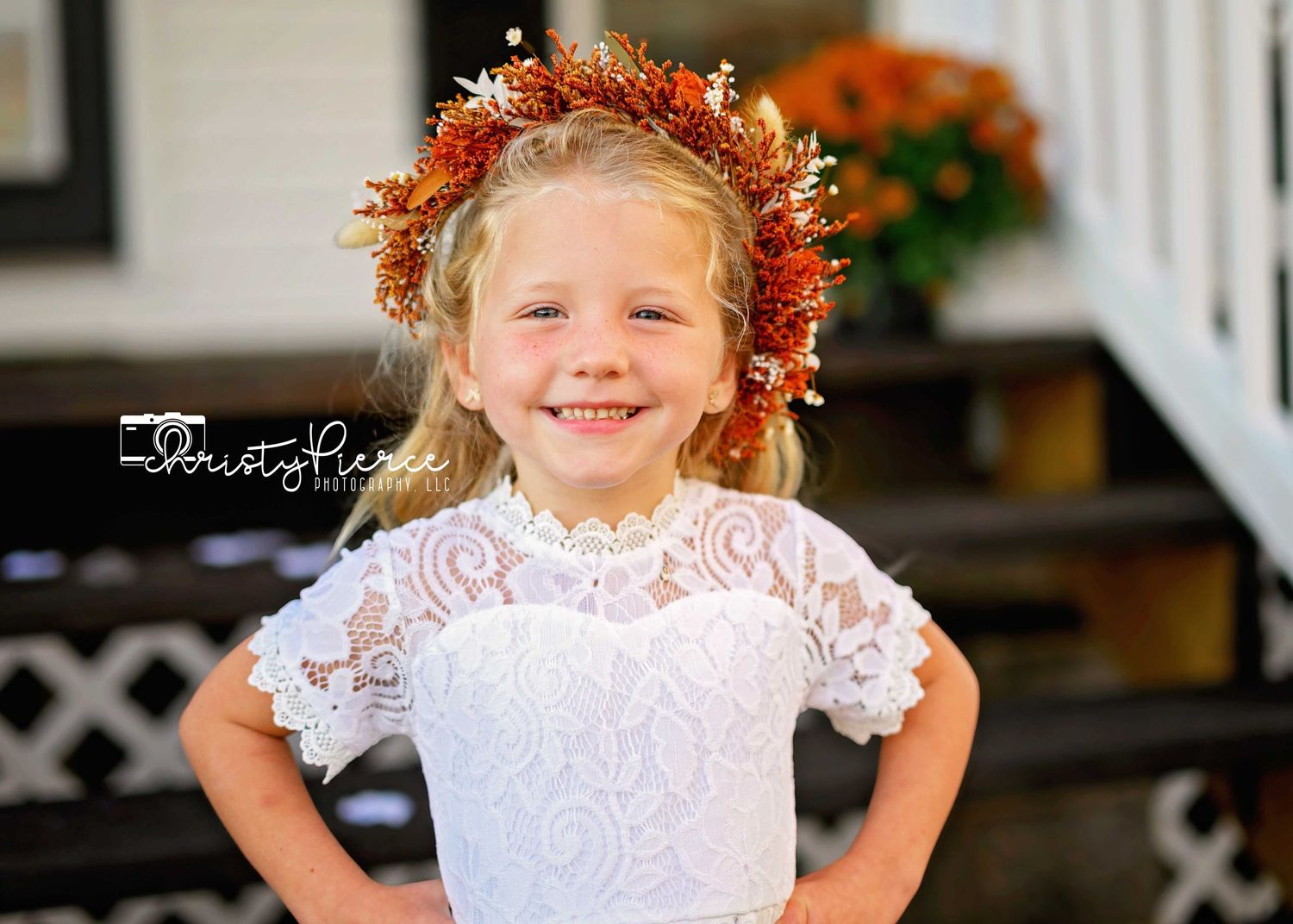 A young child with curly red hair wearing a white lace dress poses outdoors on a porch.