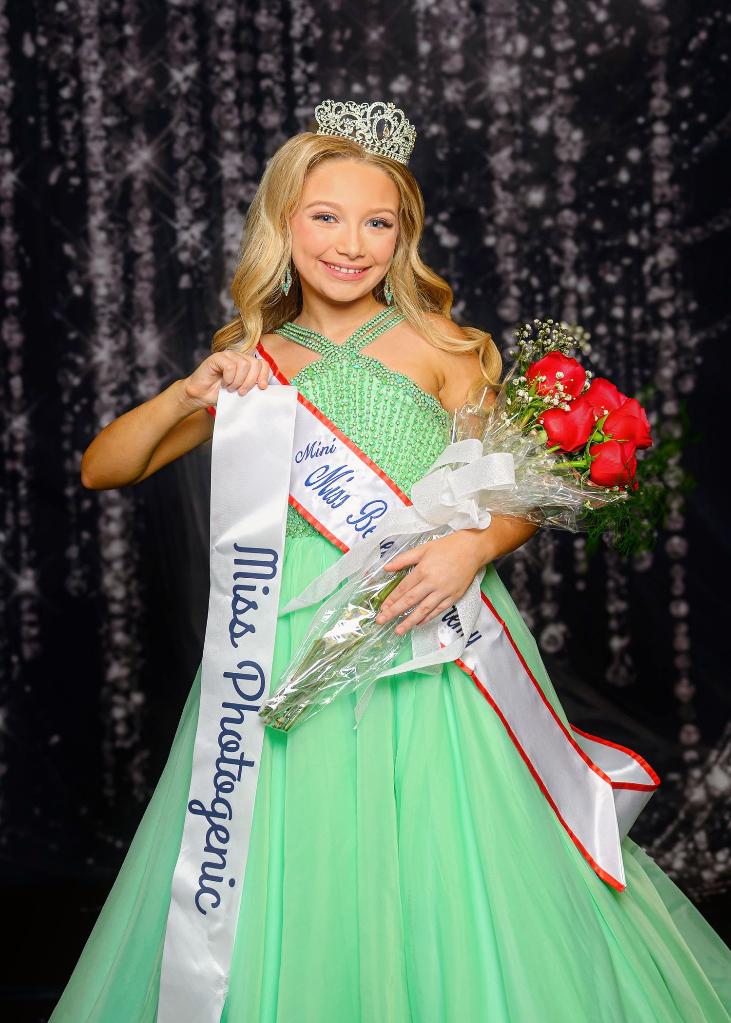 A pageant contestant in a mint green ballgown poses with flowers and a crown during a competition event.
