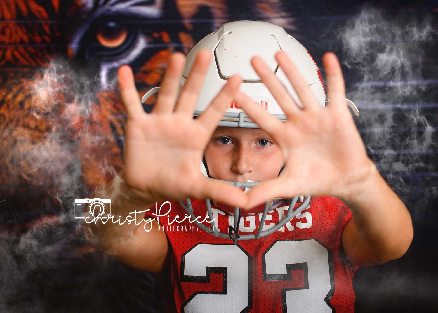 Boy in red football jersey #23 poses with hands framing face against tiger mural backdrop.