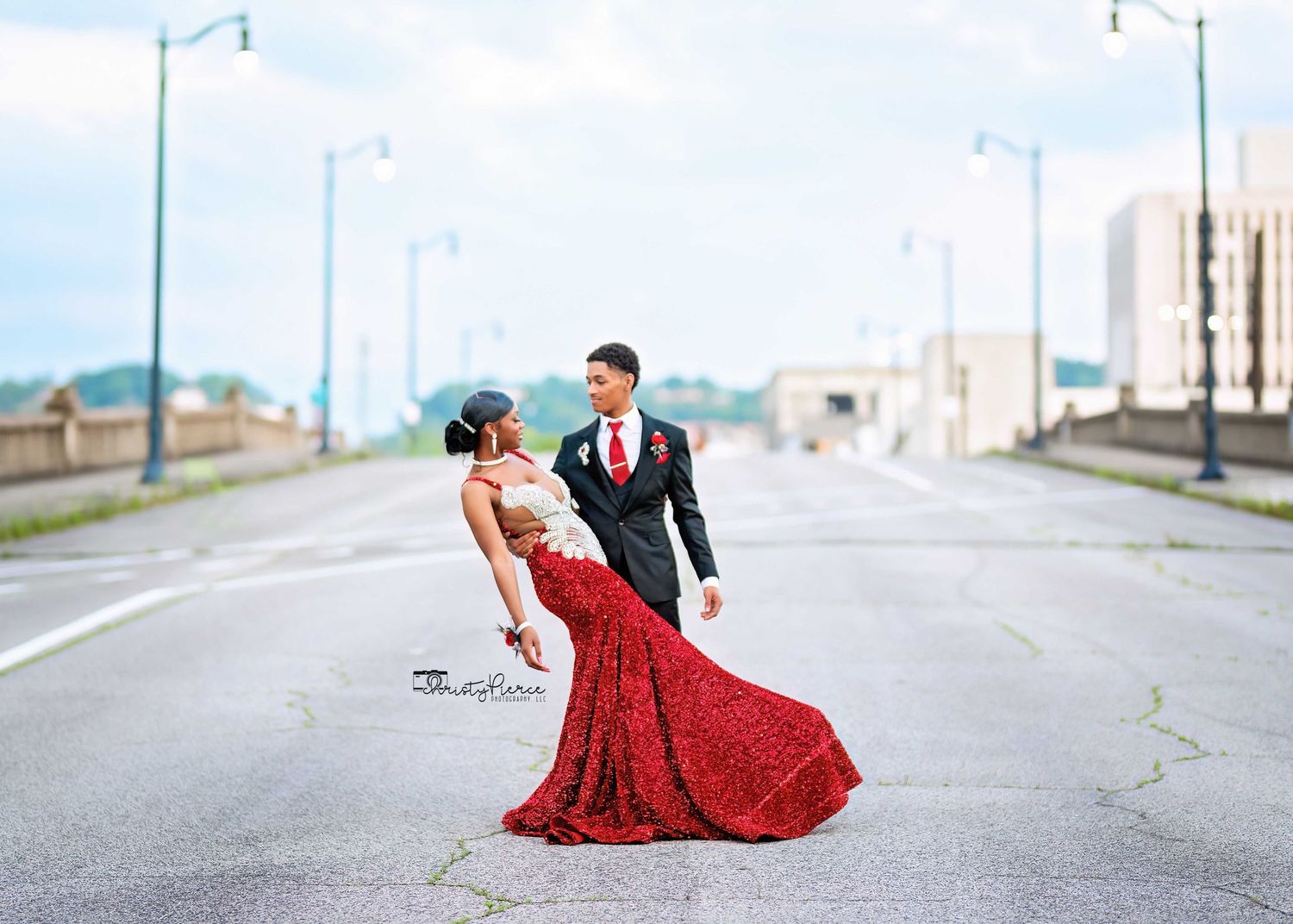 A couple in formal attire poses on an empty street, with the woman wearing a flowing red gown.
