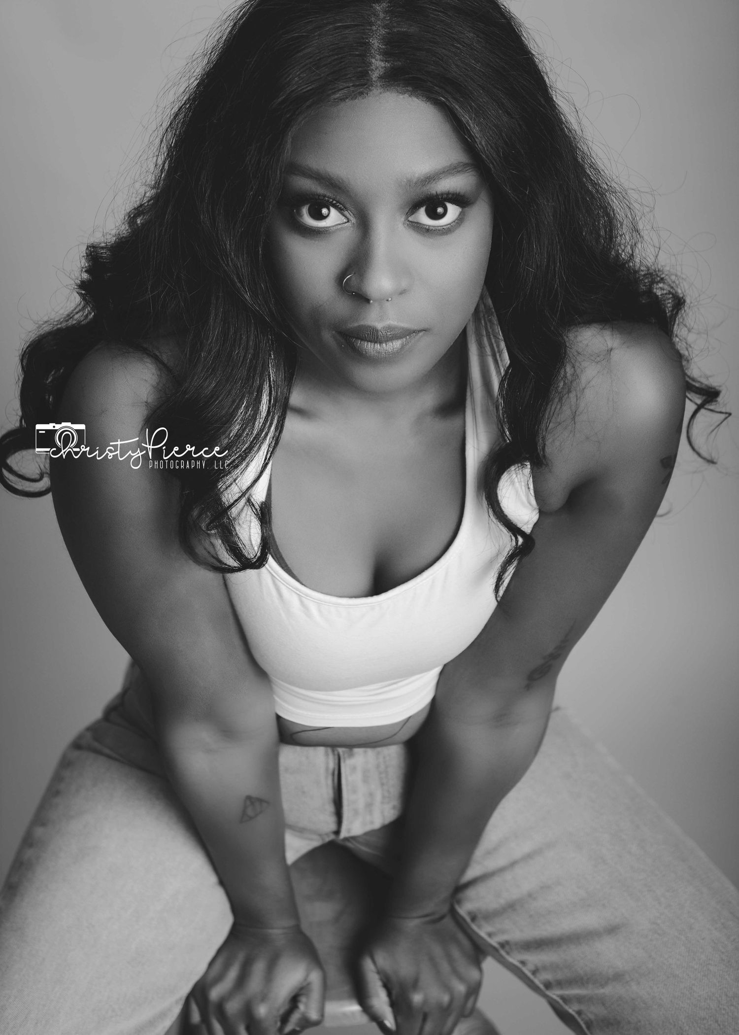 A black and white portrait of a woman in a white tank top posing with long dark hair against a light background.