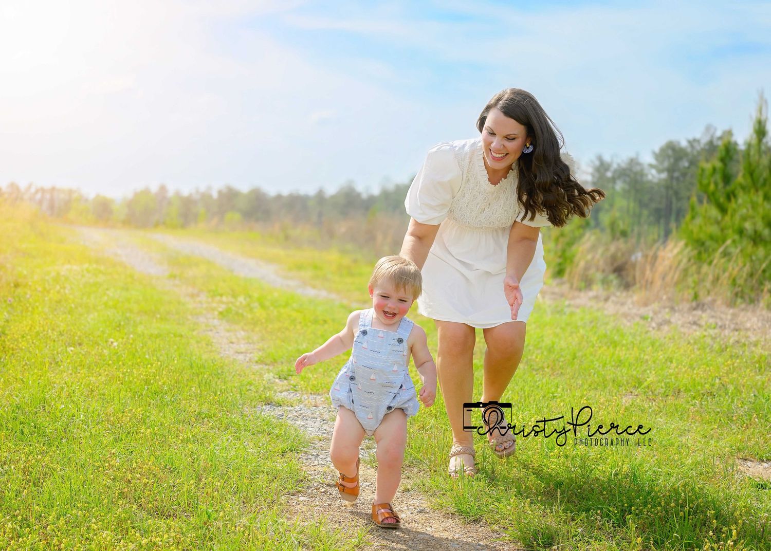 Two people walking together along a sunny dirt path through a grassy field with trees in the background.
