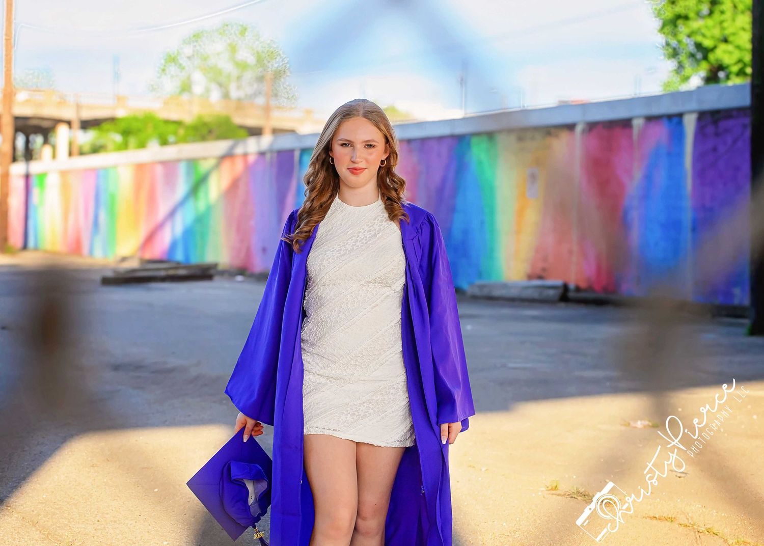 A young woman in a white dress and purple graduation gown poses in front of a colorful rainbow mural outdoors.