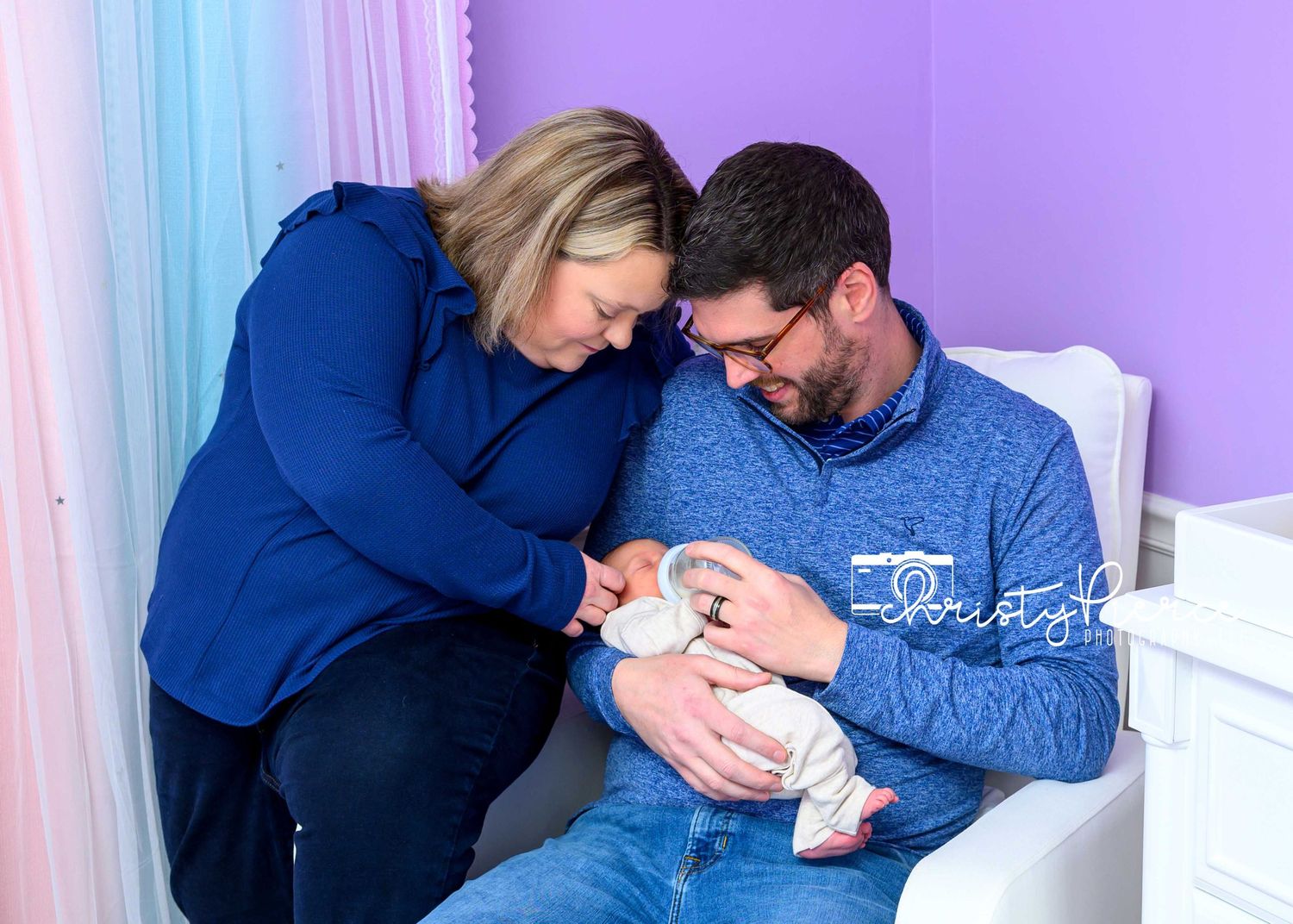 A tender family moment as parents in blue sweaters cuddle their newborn baby in a nursery setting.