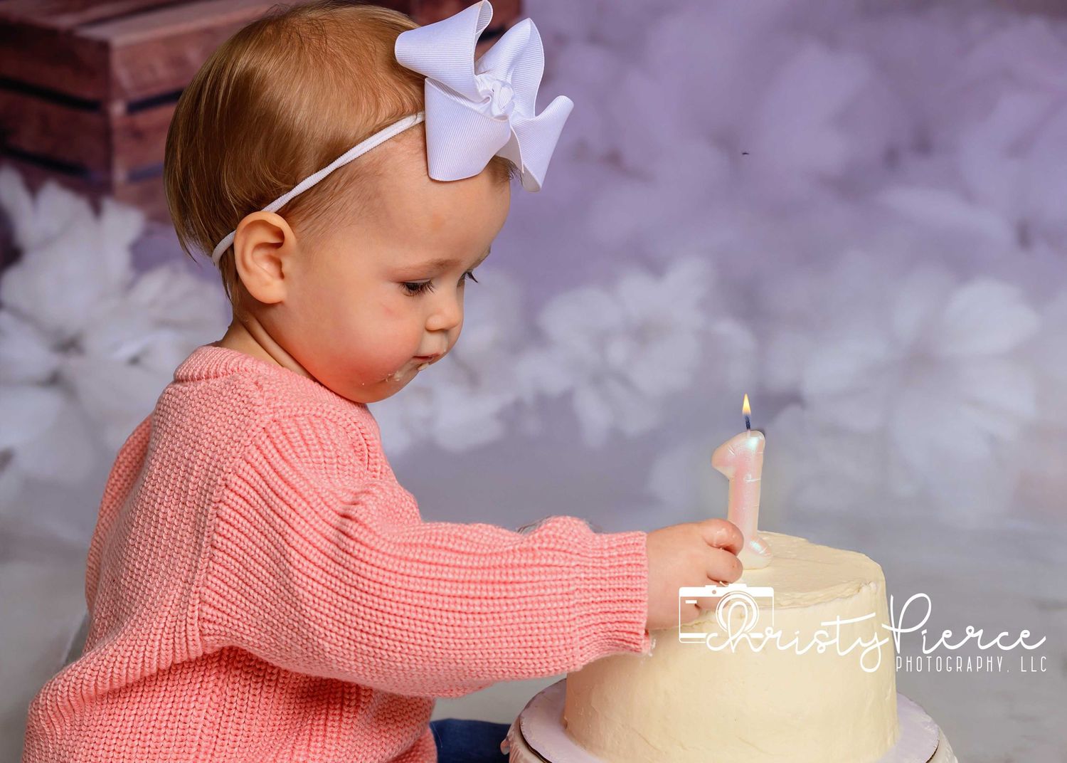A toddler in a pink sweater reaches toward a birthday cake with a lit candle.