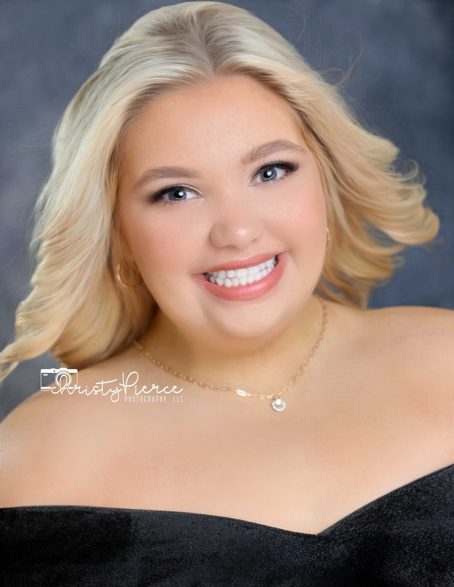 Professional portrait of someone in a black off-shoulder top with blonde hair and bright smile against a studio background.