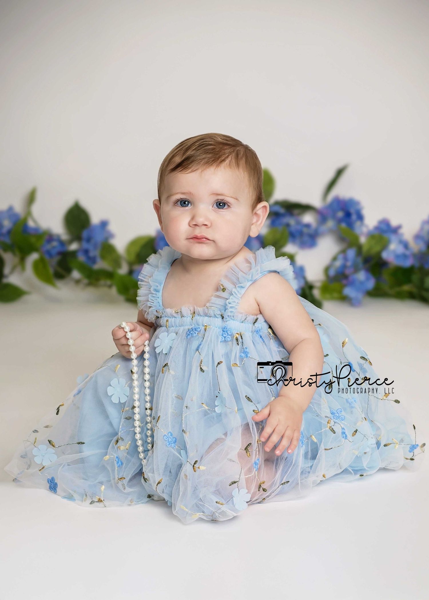Baby in light blue tulle dress poses on white backdrop with delicate blue flowers and pearl necklace decorations.