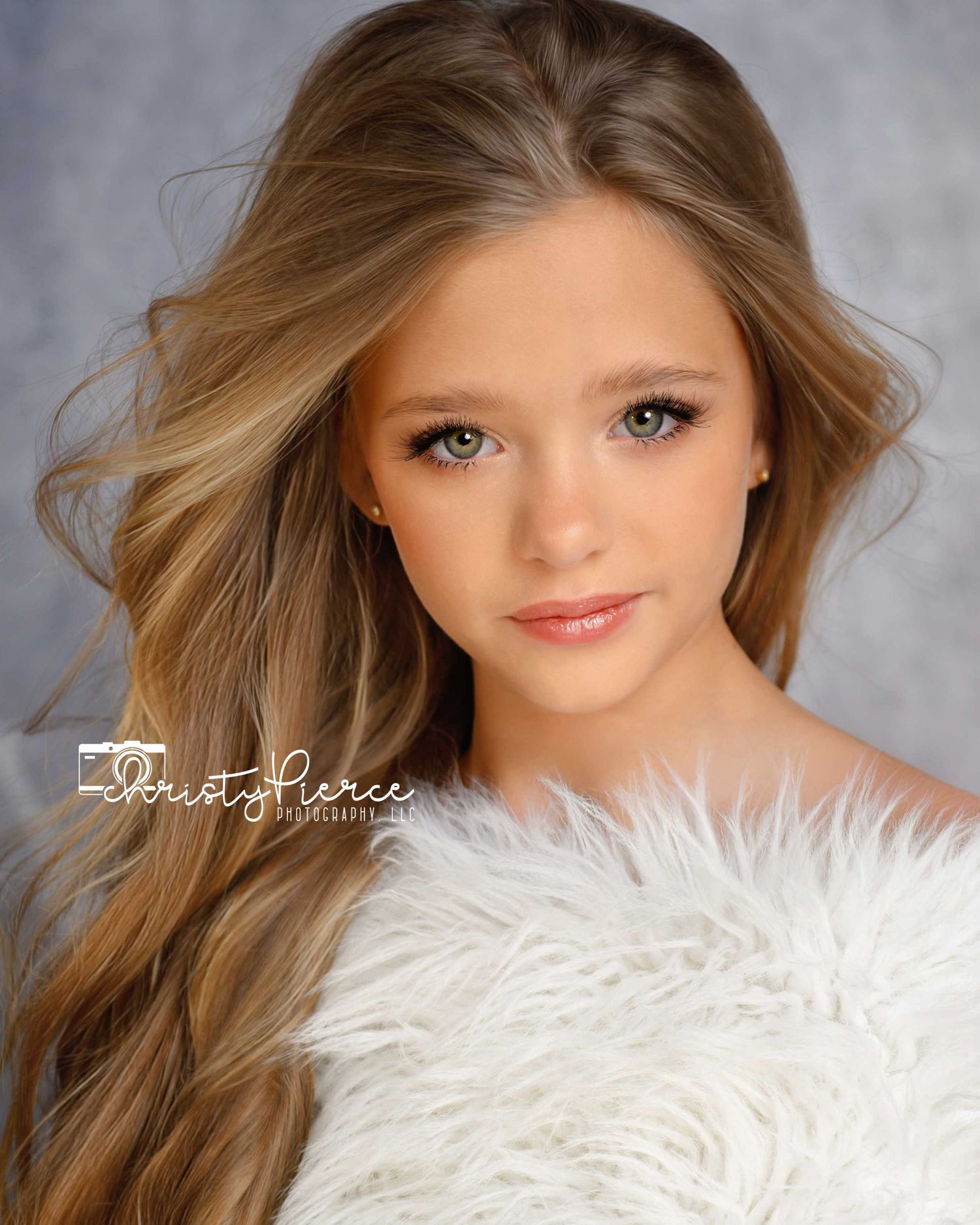 Portrait photograph featuring long brown hair and white feathered accents against a gray studio backdrop.