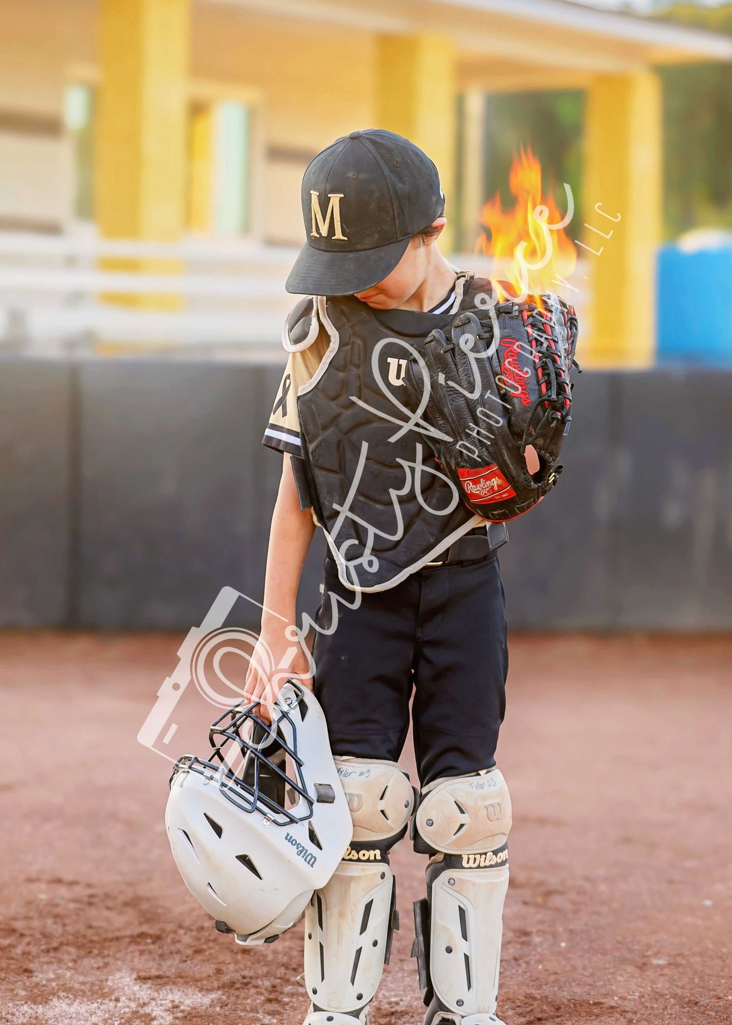 Young baseball catcher in dark uniform holds helmet and glove with leg guards at a baseball field.