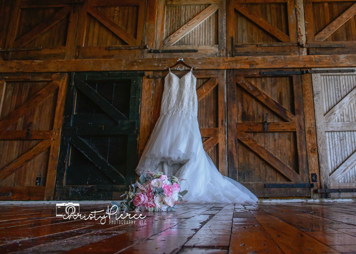 A elegant white wedding dress hangs on rustic barn doors with a pink bridal bouquet on the floor.