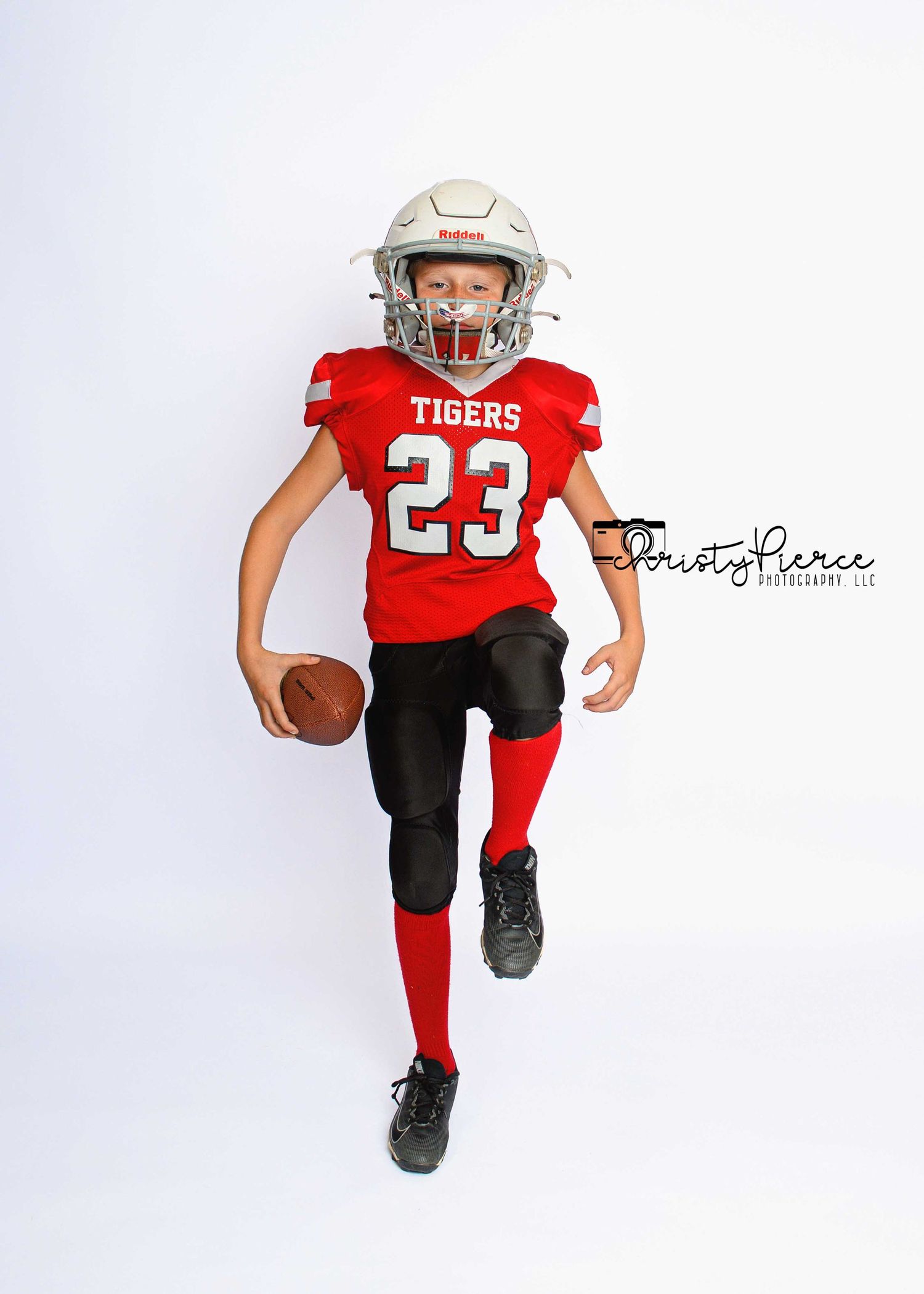 Young football player in red Tigers jersey number 23 running with ball in full uniform and helmet.
