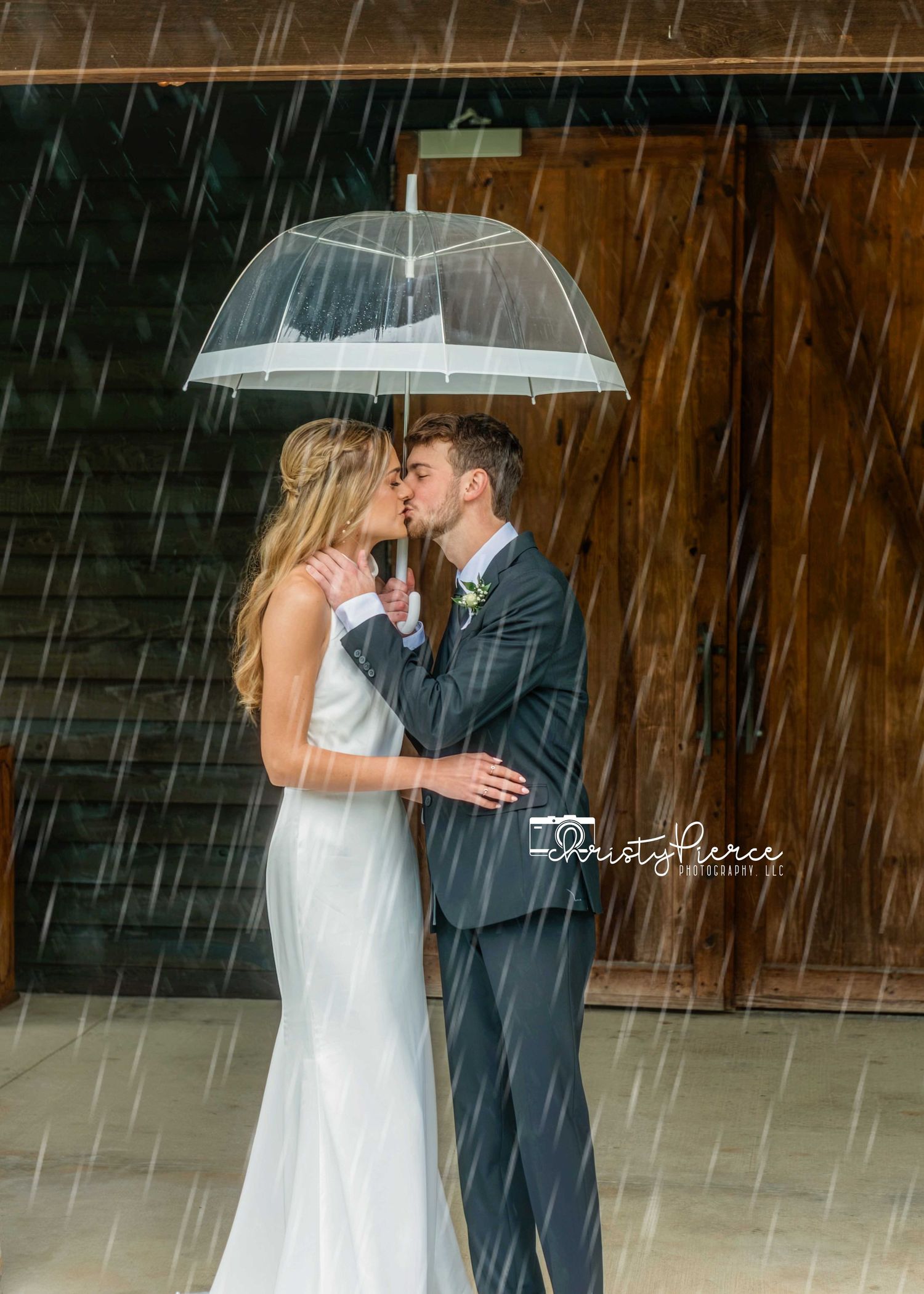 A romantic wedding couple shares an intimate moment under a clear umbrella in the rain against a rustic wooden backdrop.