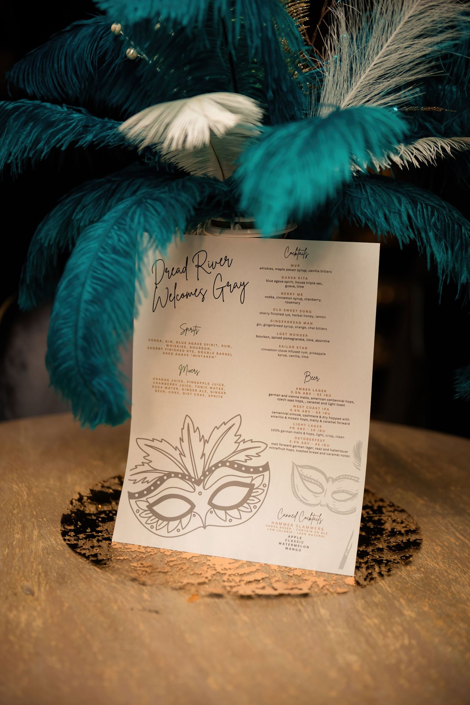 A masquerade party menu with a decorative mask design sits propped against teal feather plumes on a table.