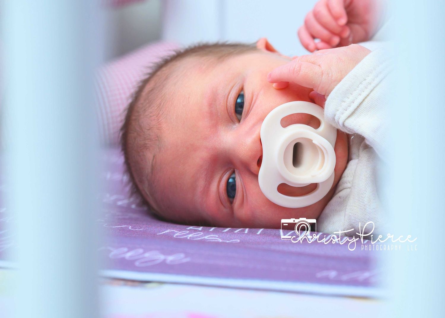 A baby with a pacifier rests peacefully on soft pink and purple bedding.