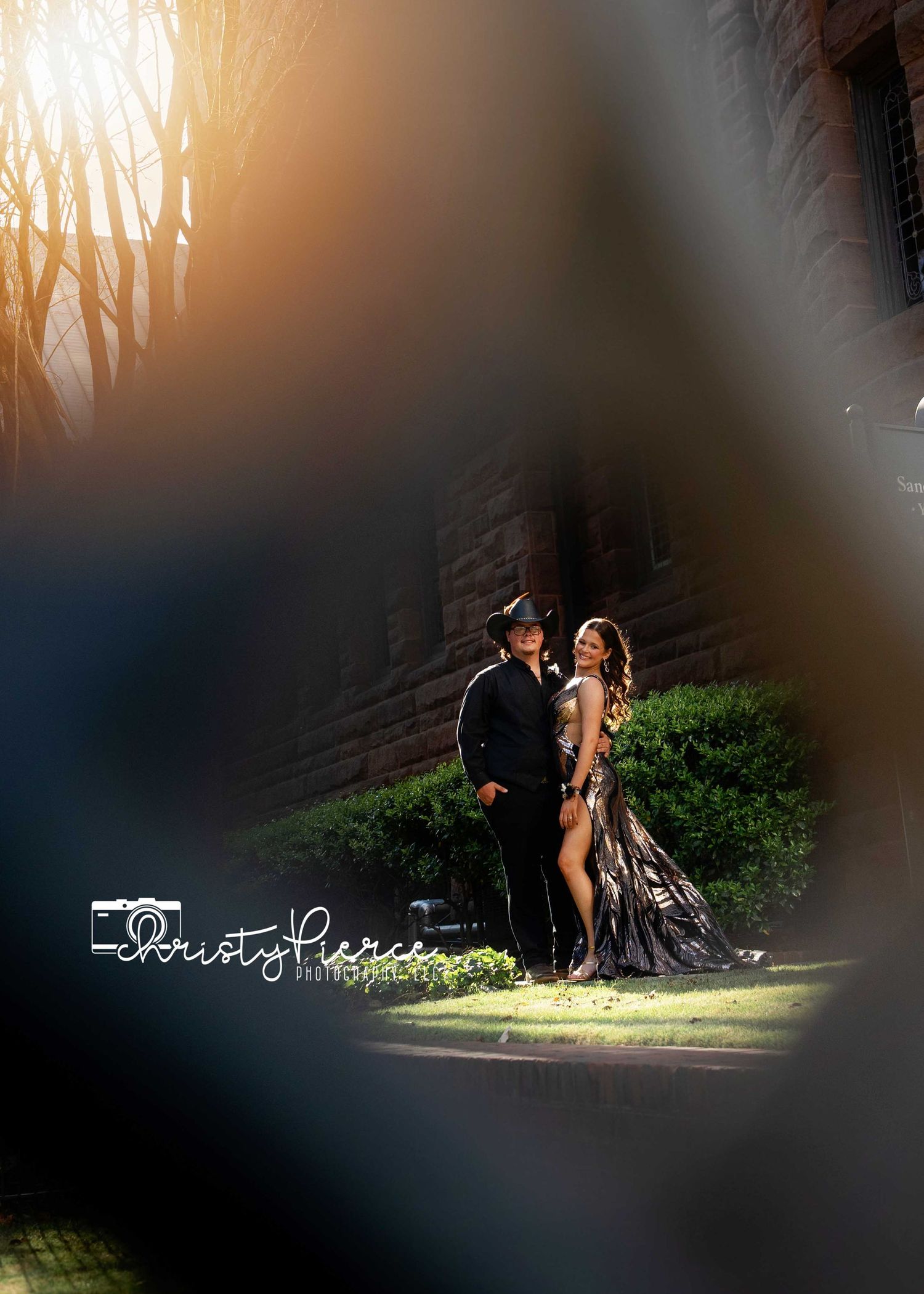 Couple poses elegantly outdoors, man in black suit and woman in dramatic dark gown, framed by bokeh foreground blur.