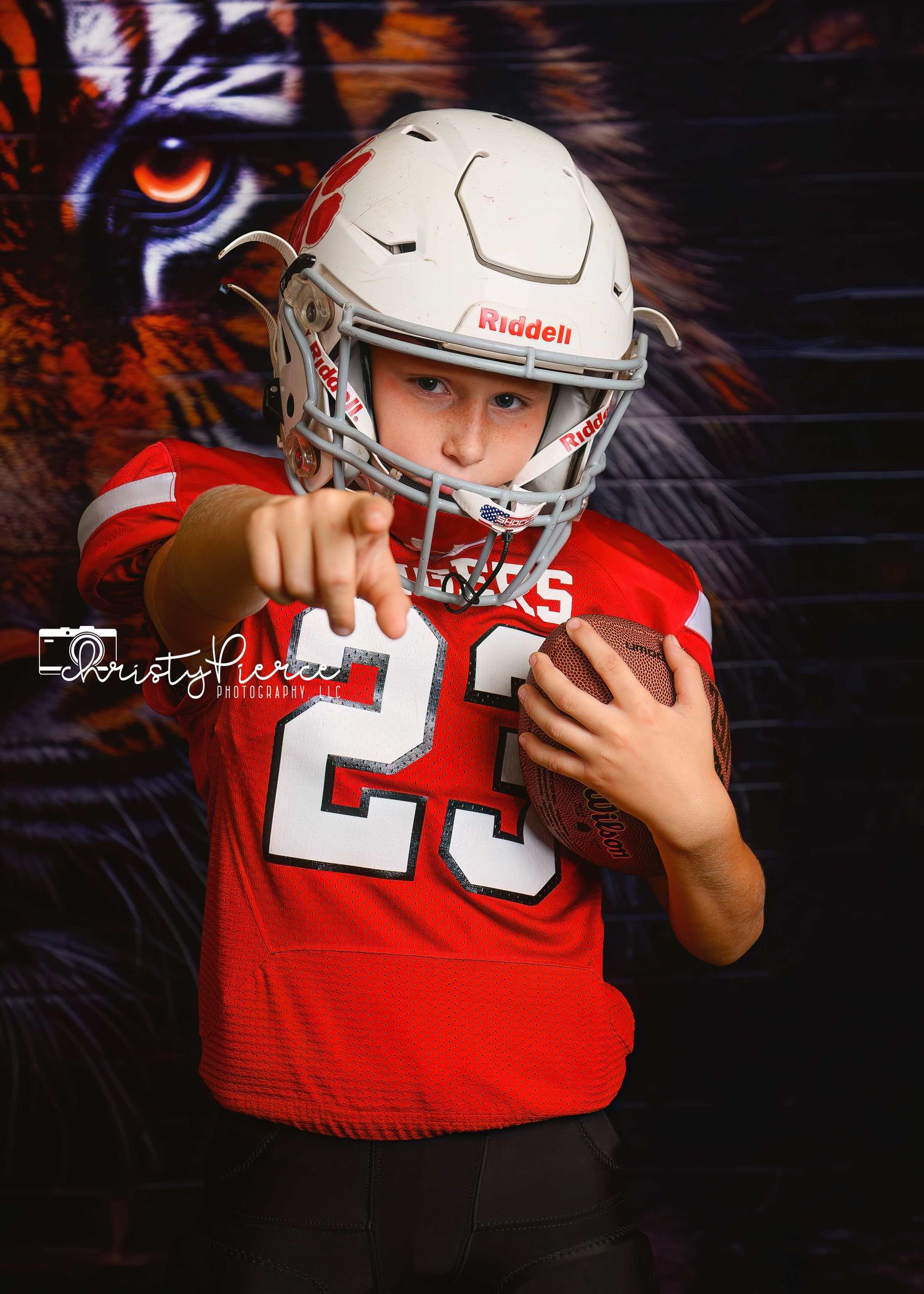 Young football player in red #23 jersey and white Riddell helmet pointing at camera holding a football with tiger backdrop.