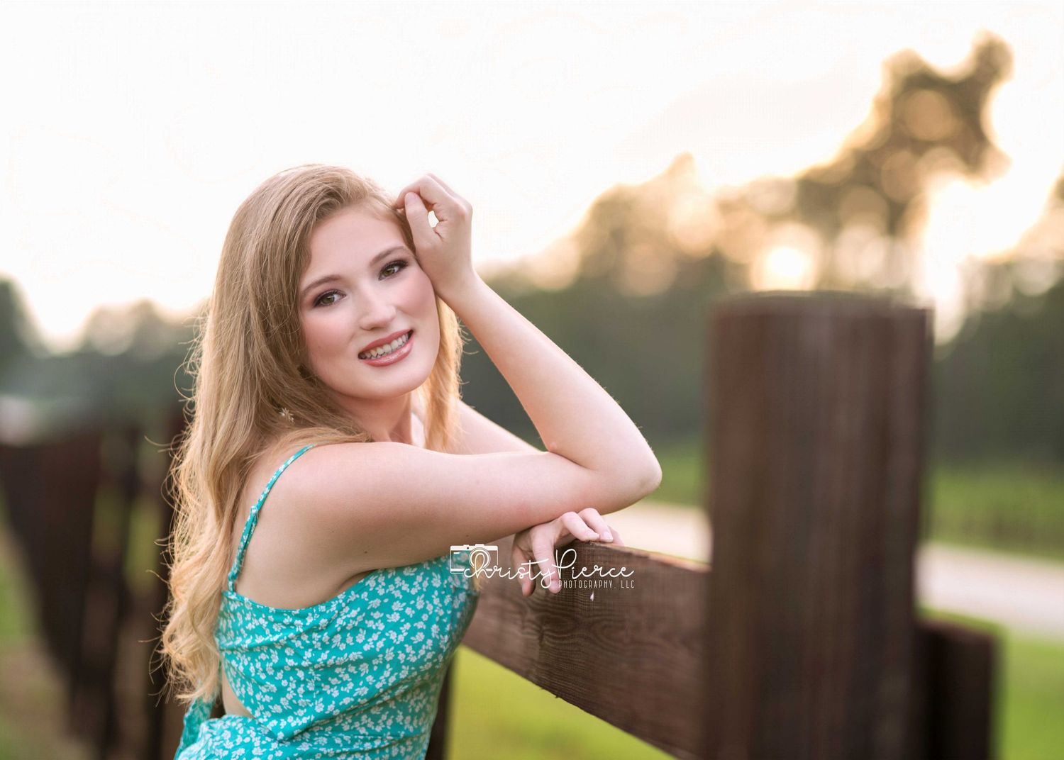 A person in a turquoise patterned dress leans on a wooden fence during golden hour sunset.